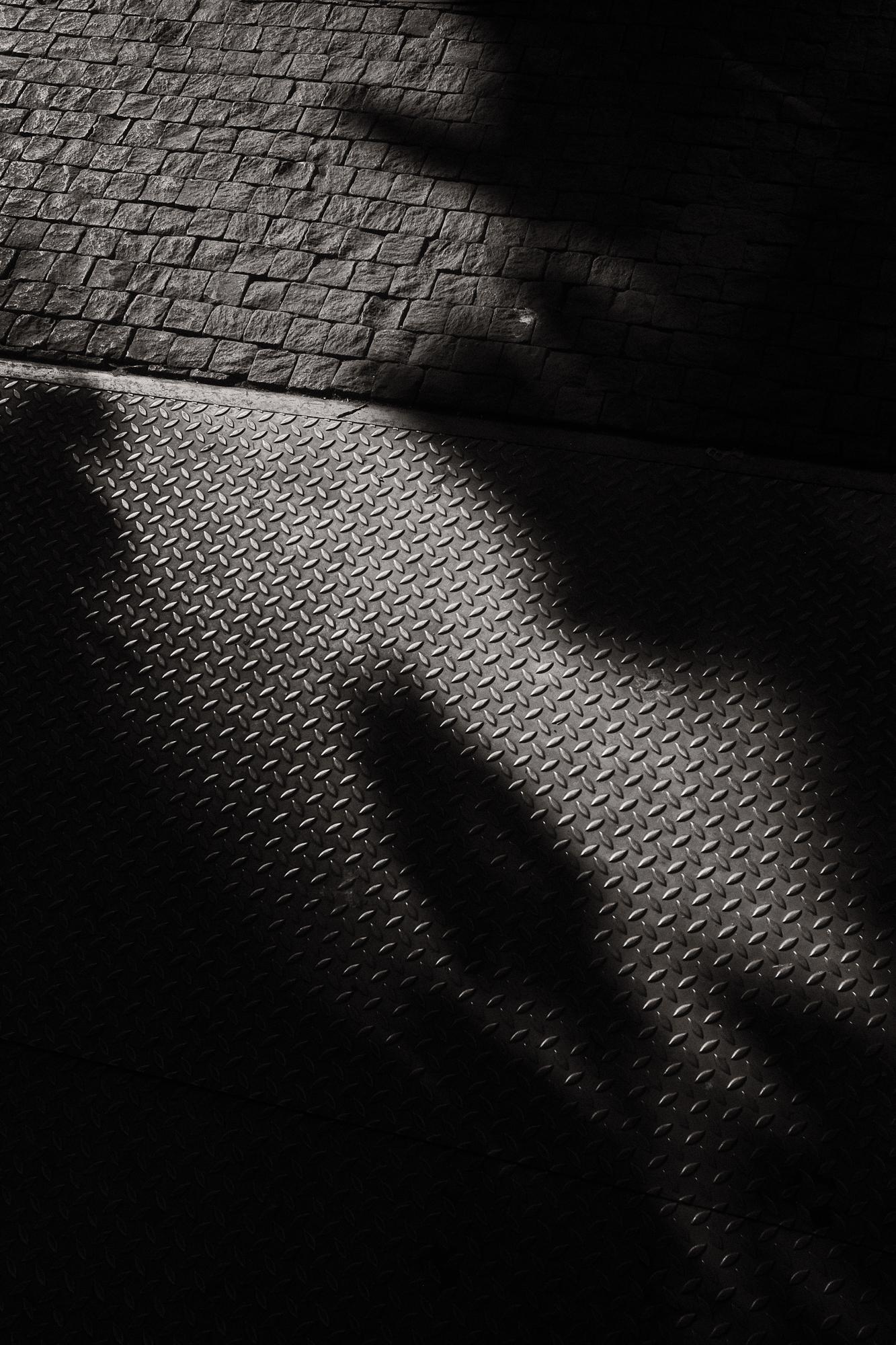 Čiernobiely záber s tieňmi na dlažobných kockách a plechu, zachytávajúci textúru a kontrasty mestského prostredia.