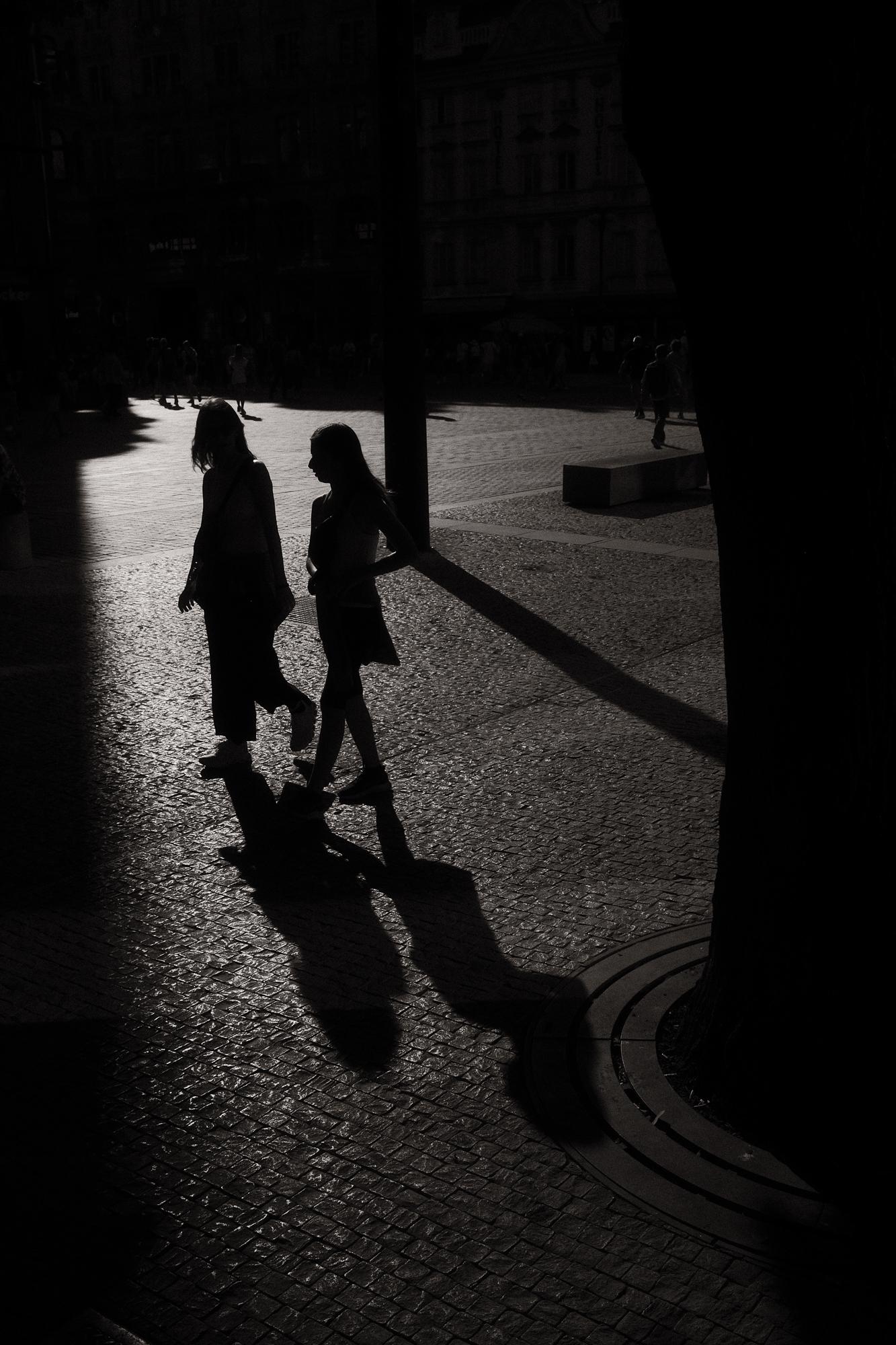 Dve osoby kráčajú po dláždenej ulici, osvetlené zapadajúcim slnkom, ich dlhé tiene sa tiahnu po zemi, atmosféra mestského večera.