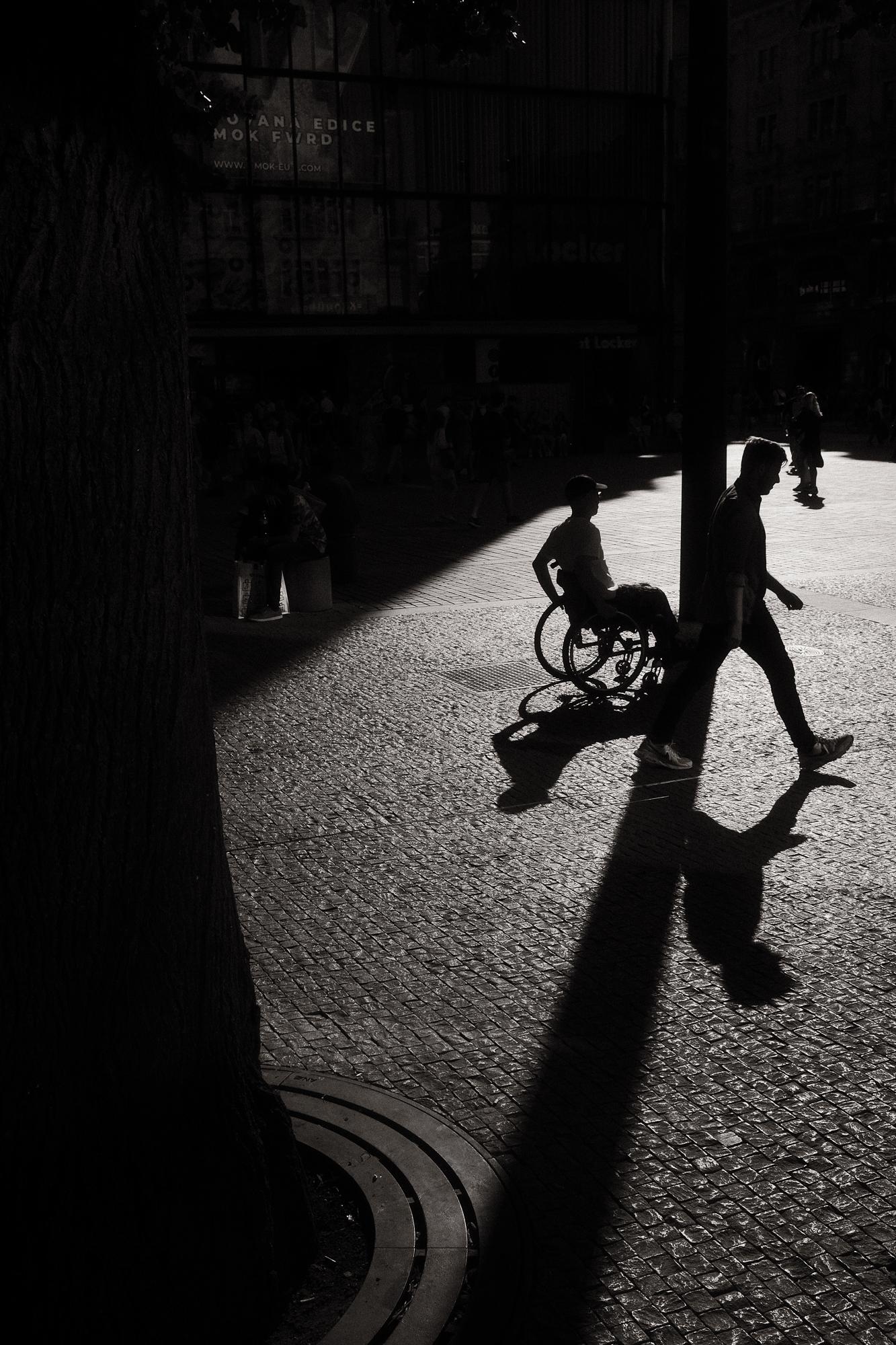 Čiernobiela fotografia ulice zachytáva ľudí na námestí. Slnko v protisvetle vytvára dlhé tiene a robí z ľudí siluety.