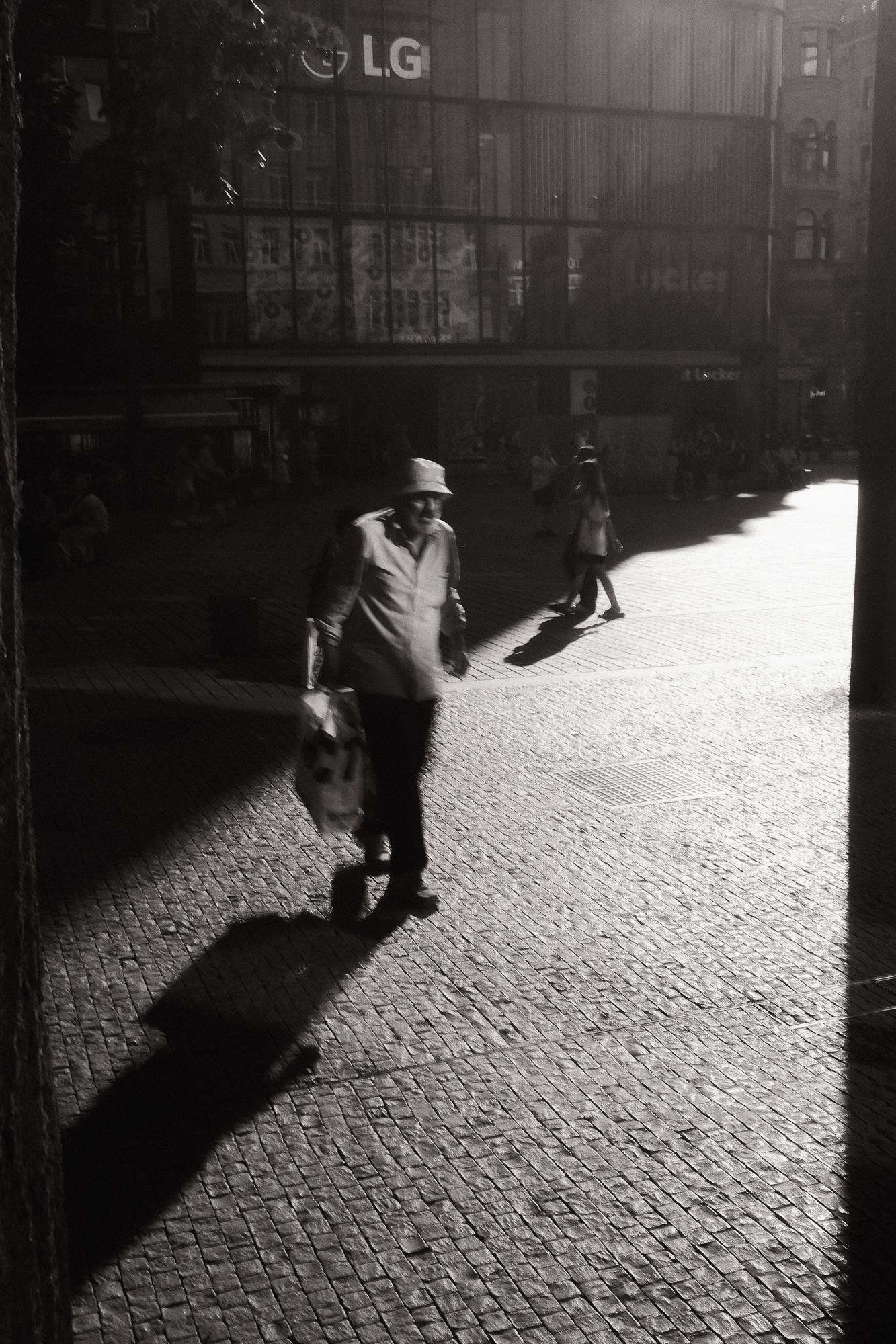 Čiernobiela fotografia muža kráčajúceho po dláždenej ulici, osvetleného prudkým svetlom s dlhým tieňom za ním. V pozadí je moderná sklenená budova.