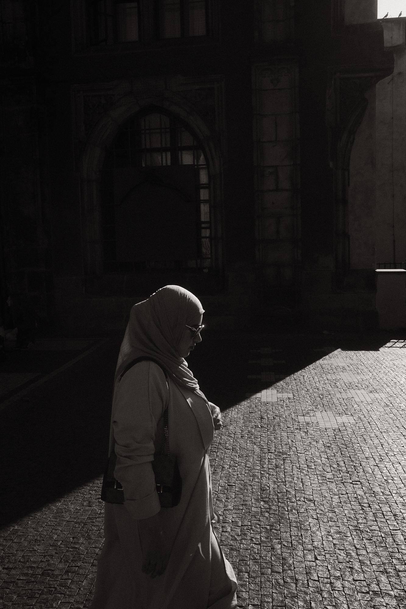 Čiernobiela fotografia zobrazujúca ženu prechádzajúcu cez historickú dlaždenú ulicu.