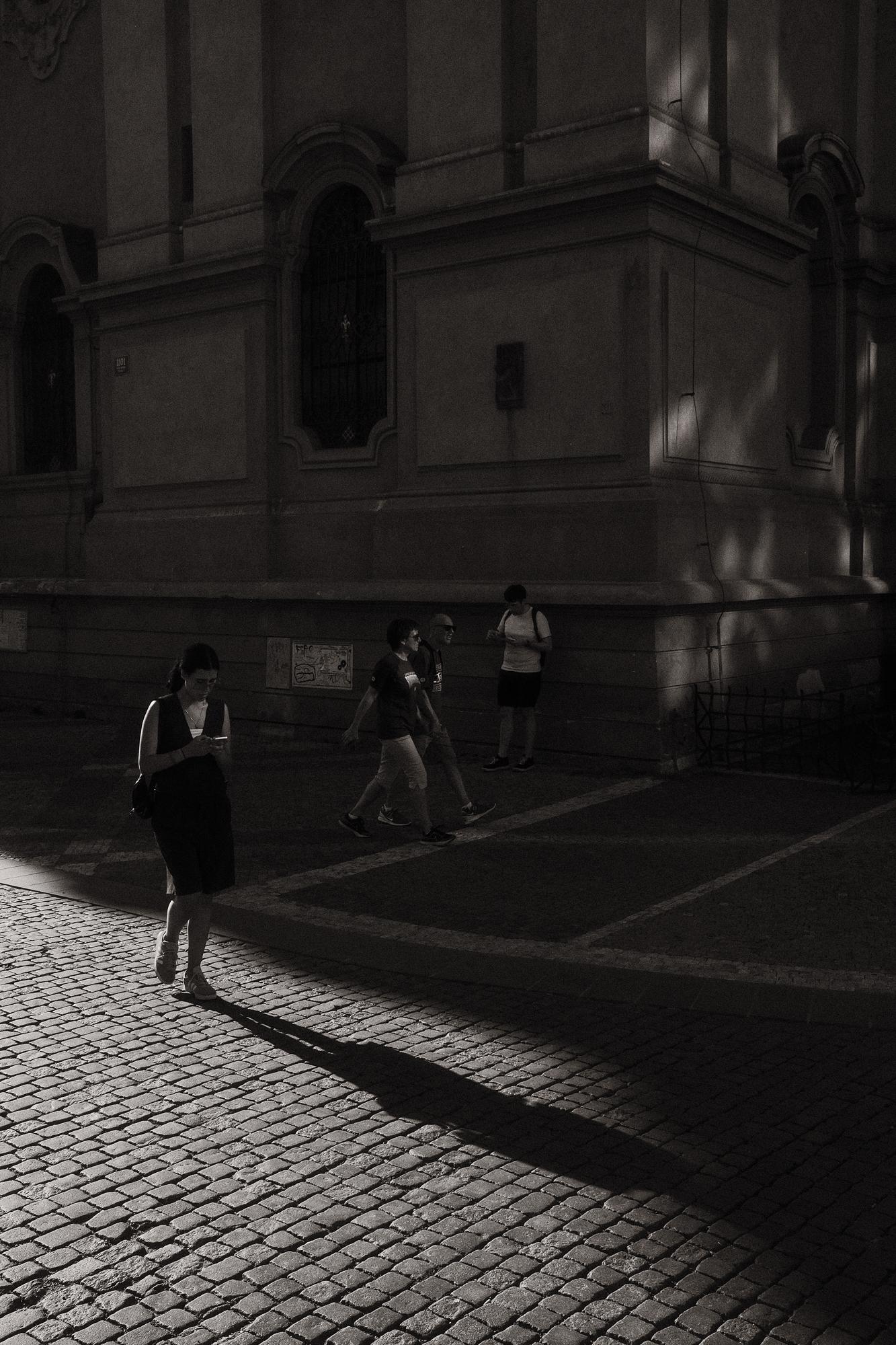 Čiernobiela fotografia ulice s ľuďmi prechádzajúcimi po dláždenom chodníku. Svetlo vytvára dramatické tiene.