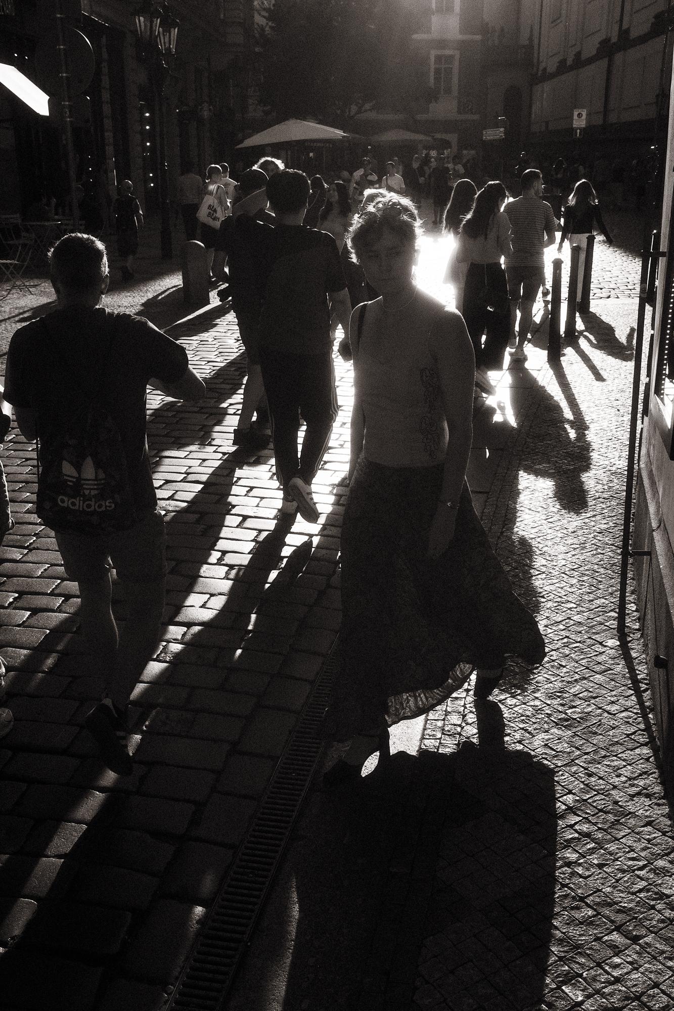 Čiernobiela fotografia ženy v slnečnom protisvetle uprostred rušnej pražskej ulice.