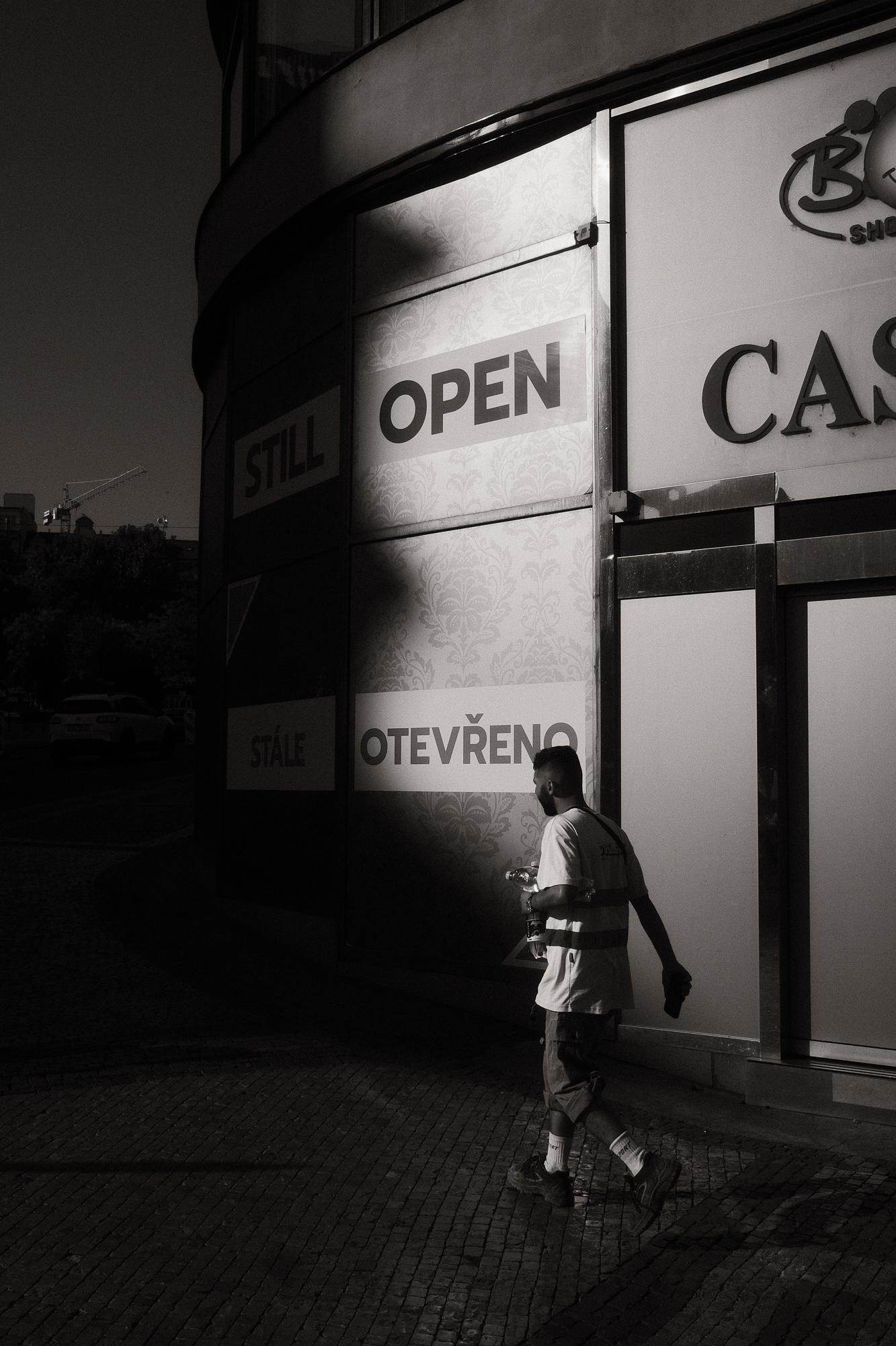 Čiernobiela fotografia zachytáva osobu kráčajúcu po dláždenej ulici pri budove kasína. Svetlo a tieň tvoria dramatický kontrast na fasáde budovy.