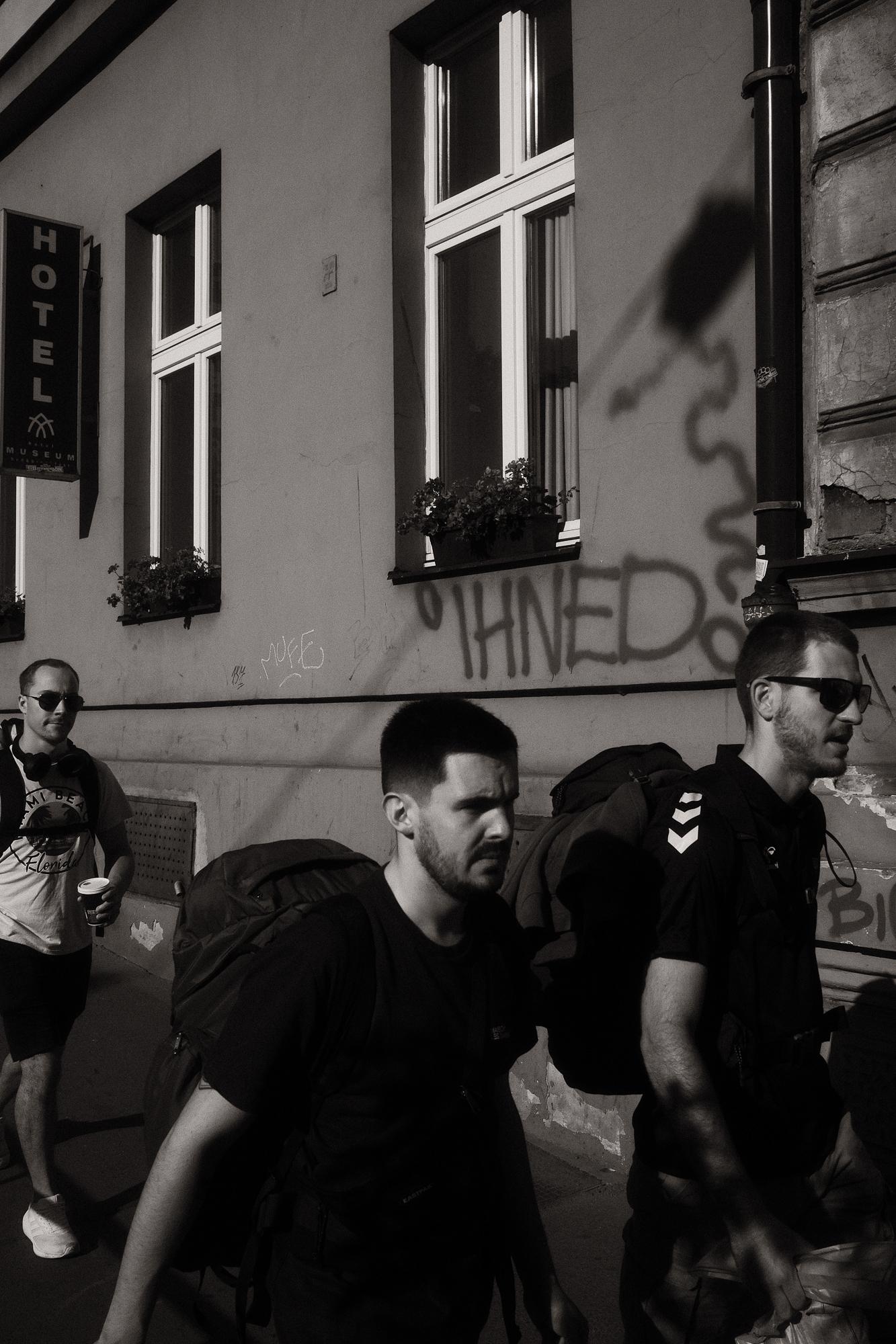 Čiernobiela fotografia troch mužov s veľkými ruksakmi kráčajúcich po chodníku. Na stene za nimi je nasprejovaný nápis "Ihned".