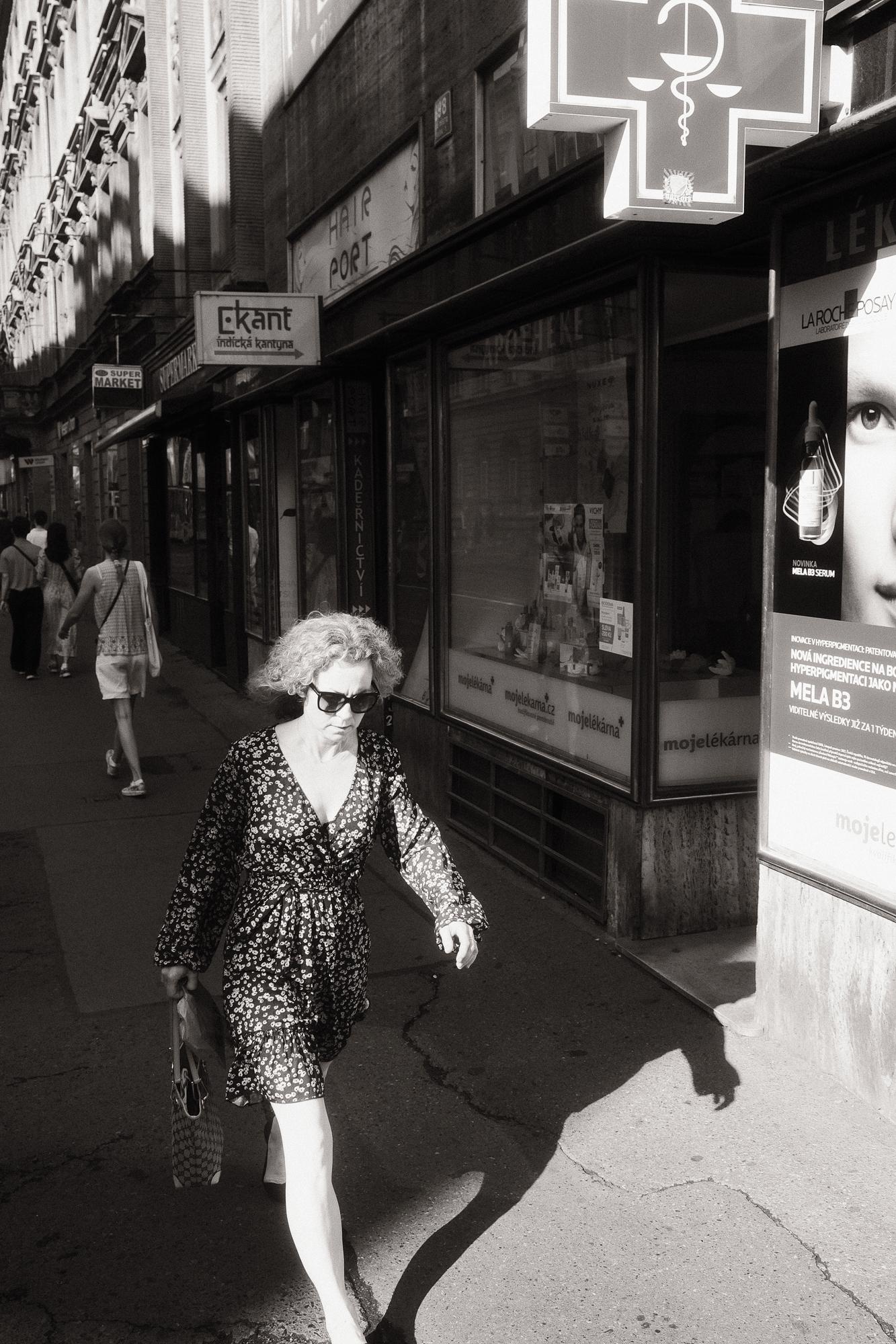 Čiernobiela fotografia ulice s ženou v kvetinových šatách, ktorá kráča po chodníku. Slnko vrhá dlhé tiene a osvetľuje časť jej tváre.