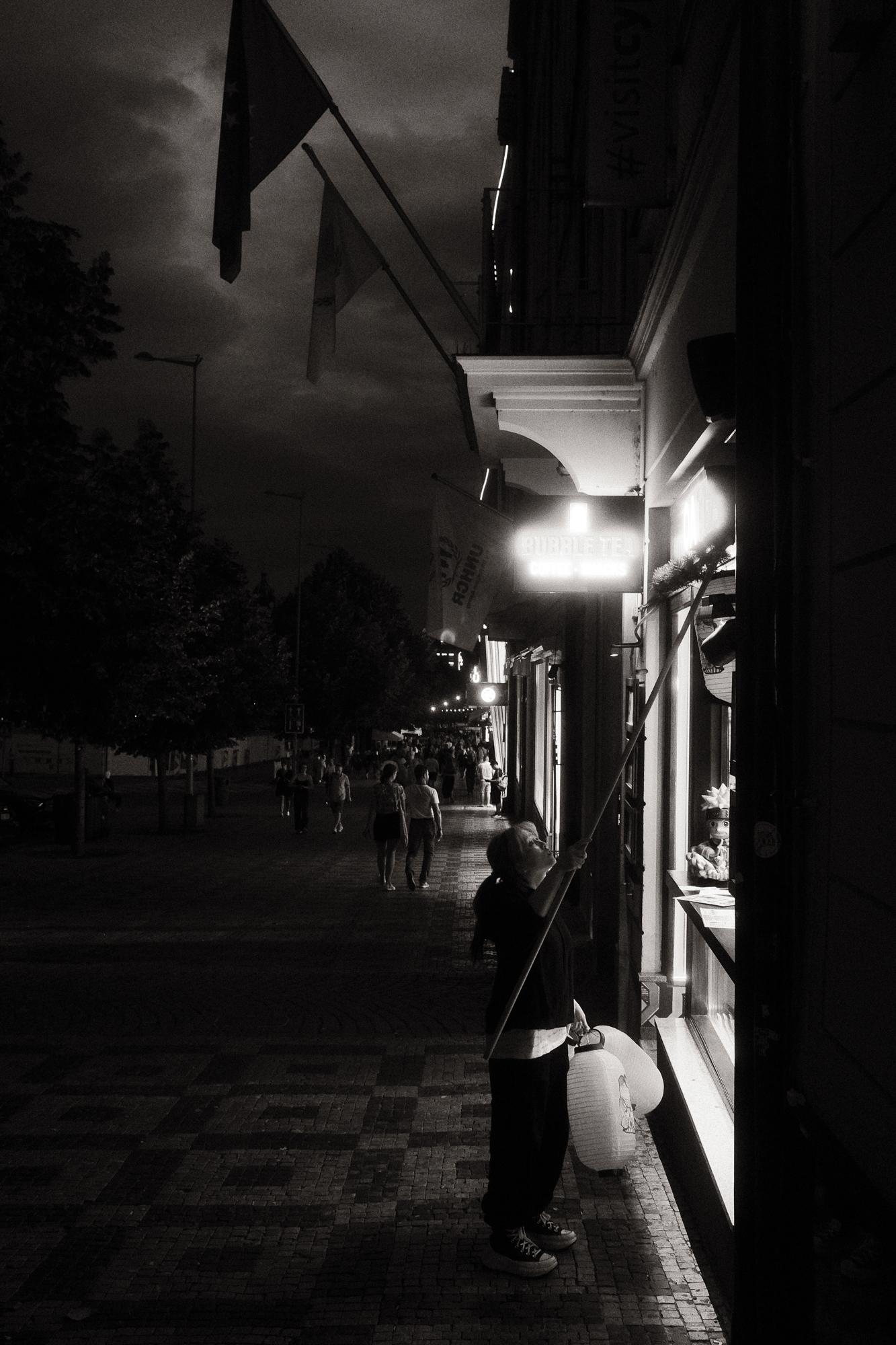 Čiernobiela fotografia ulice zachytáva ženu v noci, ktorá umýva výklad obchodu, zatiaľ čo ľudia prechádzajú po ulici.