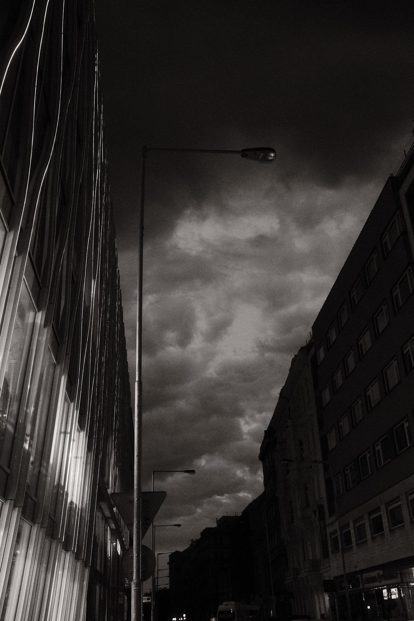 Čiernobiela fotografia večernej ulice s dramatickými oblakmi.