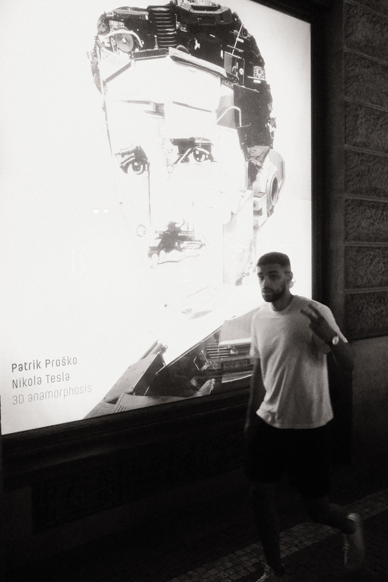 Čiernobiela fotografia muža, ktorý kráča pred veľkým plagátom Nikola Tesly.
