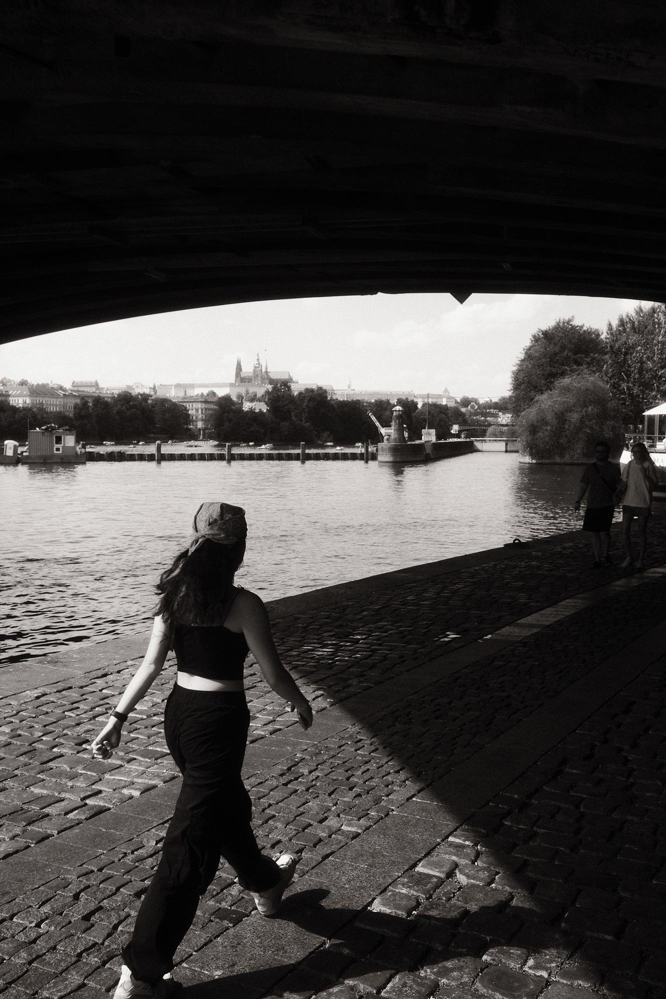 Čiernobiela fotografia ženy kráčajúcej po pražskej náplavke.