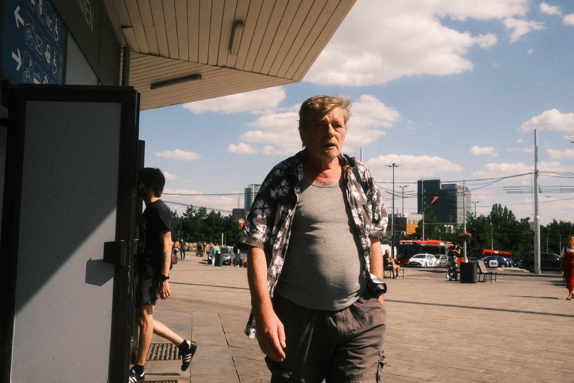 Muž v tričku a košeľi kráča po ulici pred budovou hlavnej stanice v Bratislave.