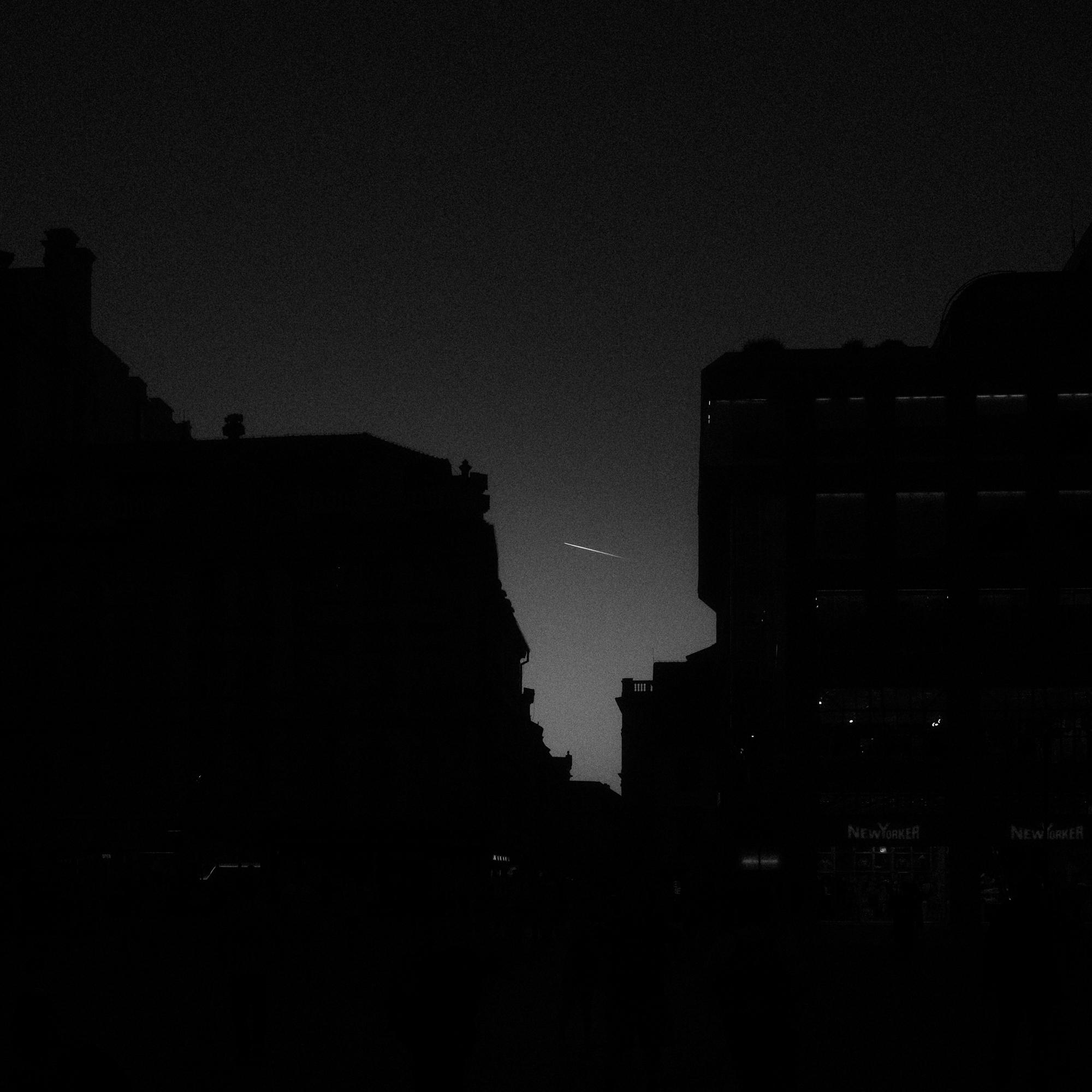 Silueta mestských budov pri súmraku s viditeľnou stopou lietadla na oblohe.