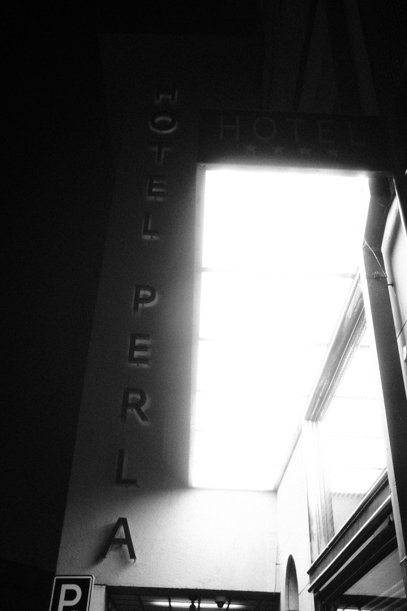 Čierno-biela fotografia hotela s nápisom PERLA na stene, vedľa svietiacej markízy.