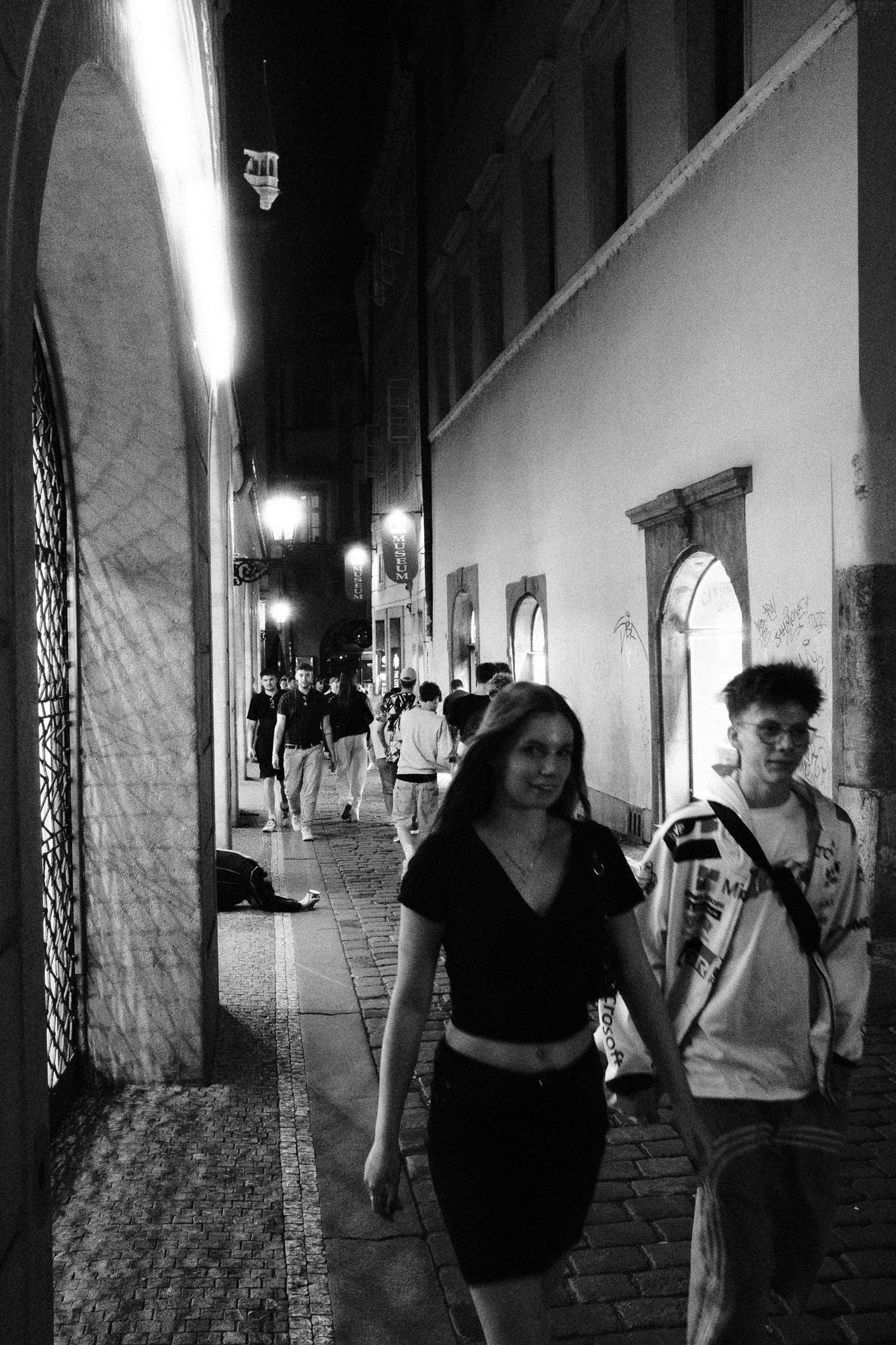 Čiernobiela fotografia ulice v noci. Na pravej strane sú dvaja ľudia kráčajúci po chodníku. V pozadí sú ďalší chodci a jasne osvetlené lampy.