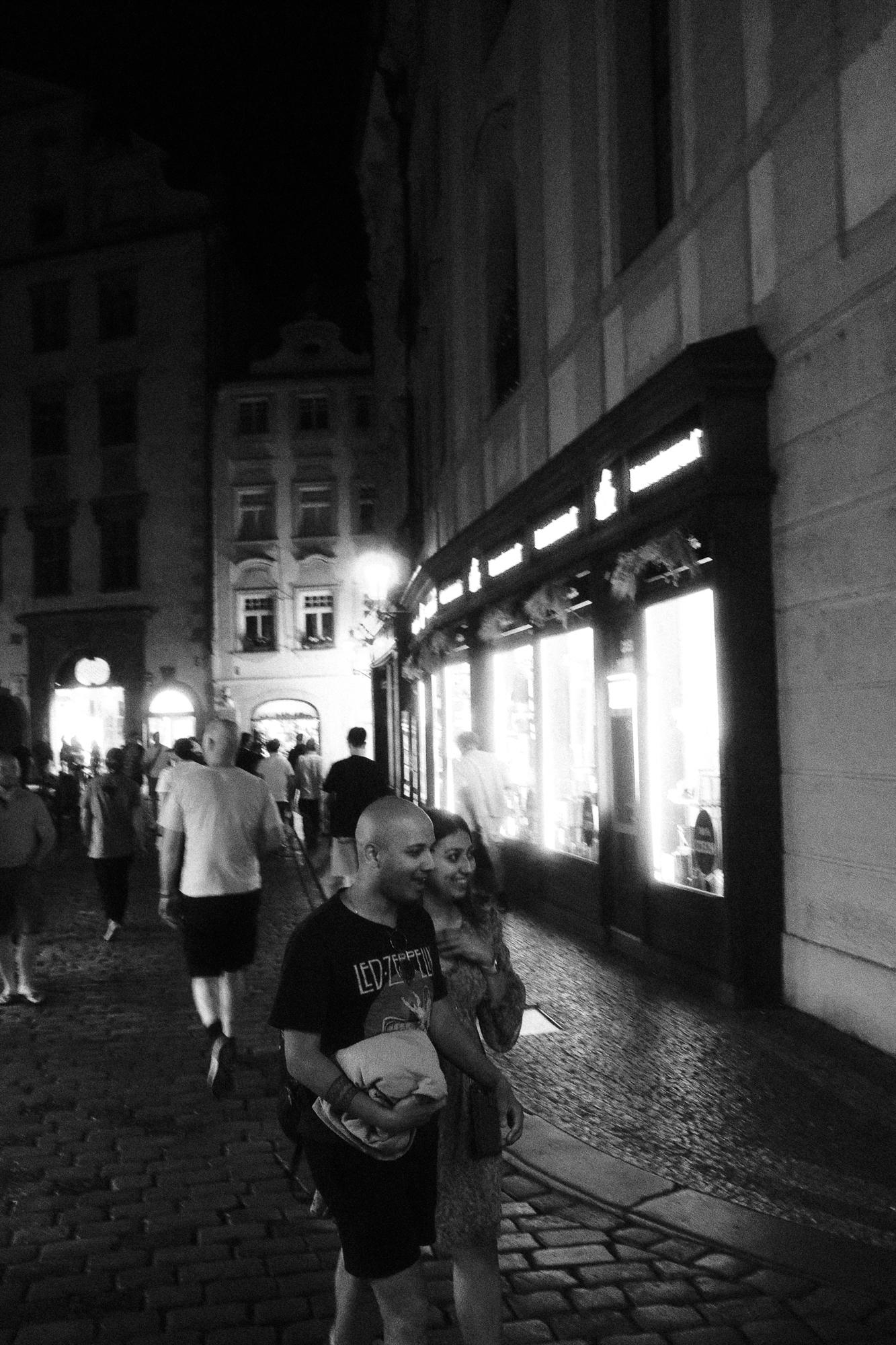 Nočná ulica s dláždeným chodníkom, dvojica nadšených turistov kráča popri osvetlenom obchode.