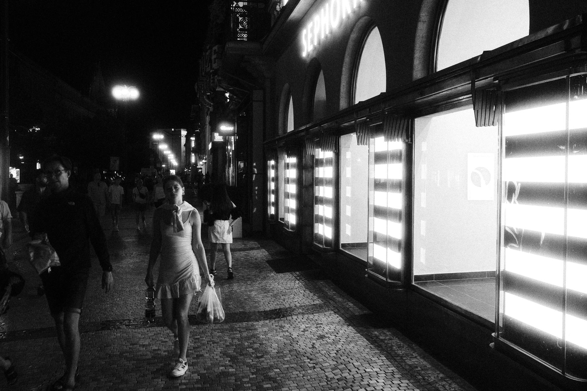 Čiernobiela fotografia ulice v noci, ľudia kráčajú popri osvetlených výkladoch na pešej zóne, dlažobné kocky na chodníku.