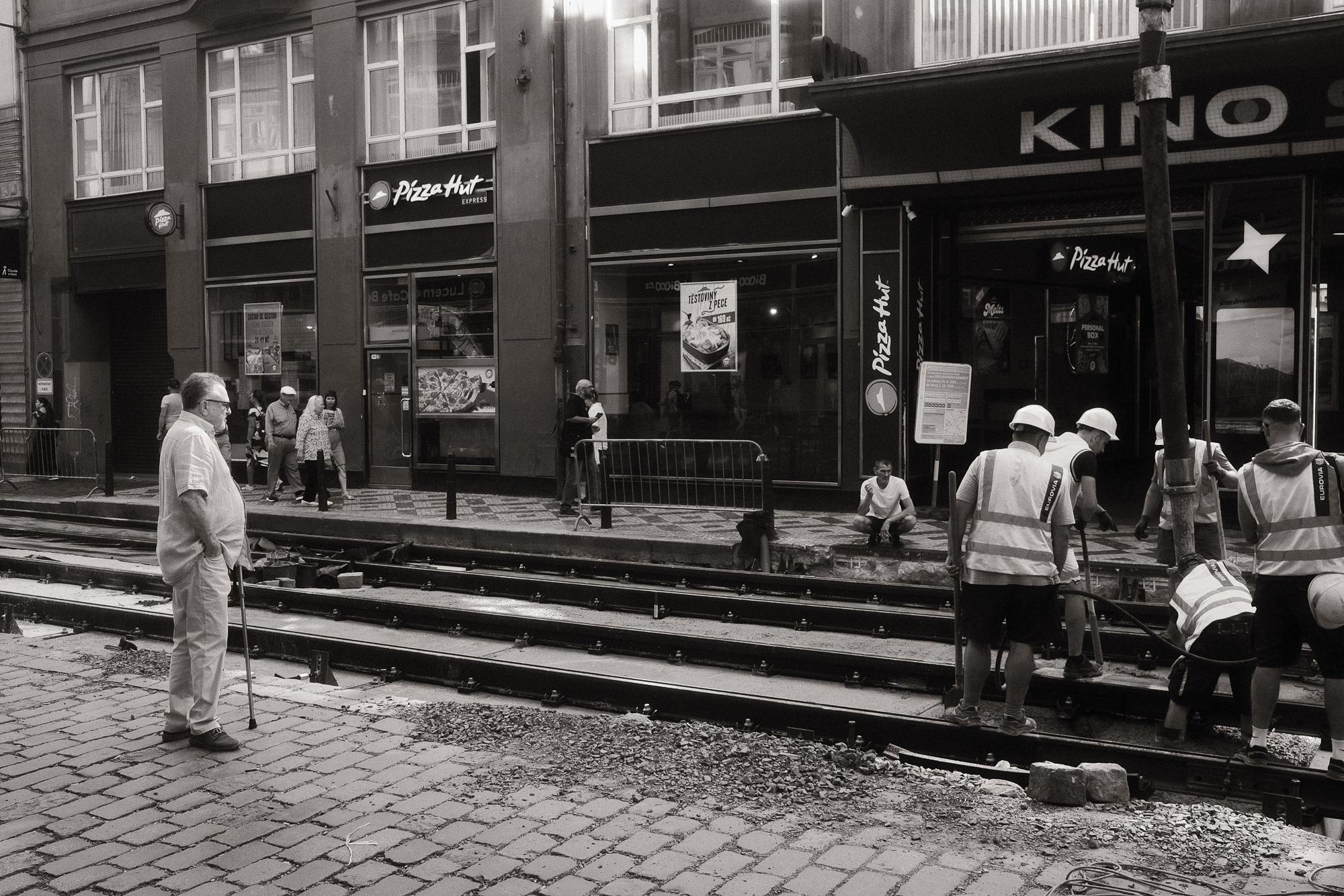 Čierno-biela fotografia mestského prostredia, kde muži v reflexných vestách pracujú na opravách koľajovej trate pred budovami s obchodmi. Starší muž s palicou sleduje prácu z opačnej strany ulice.