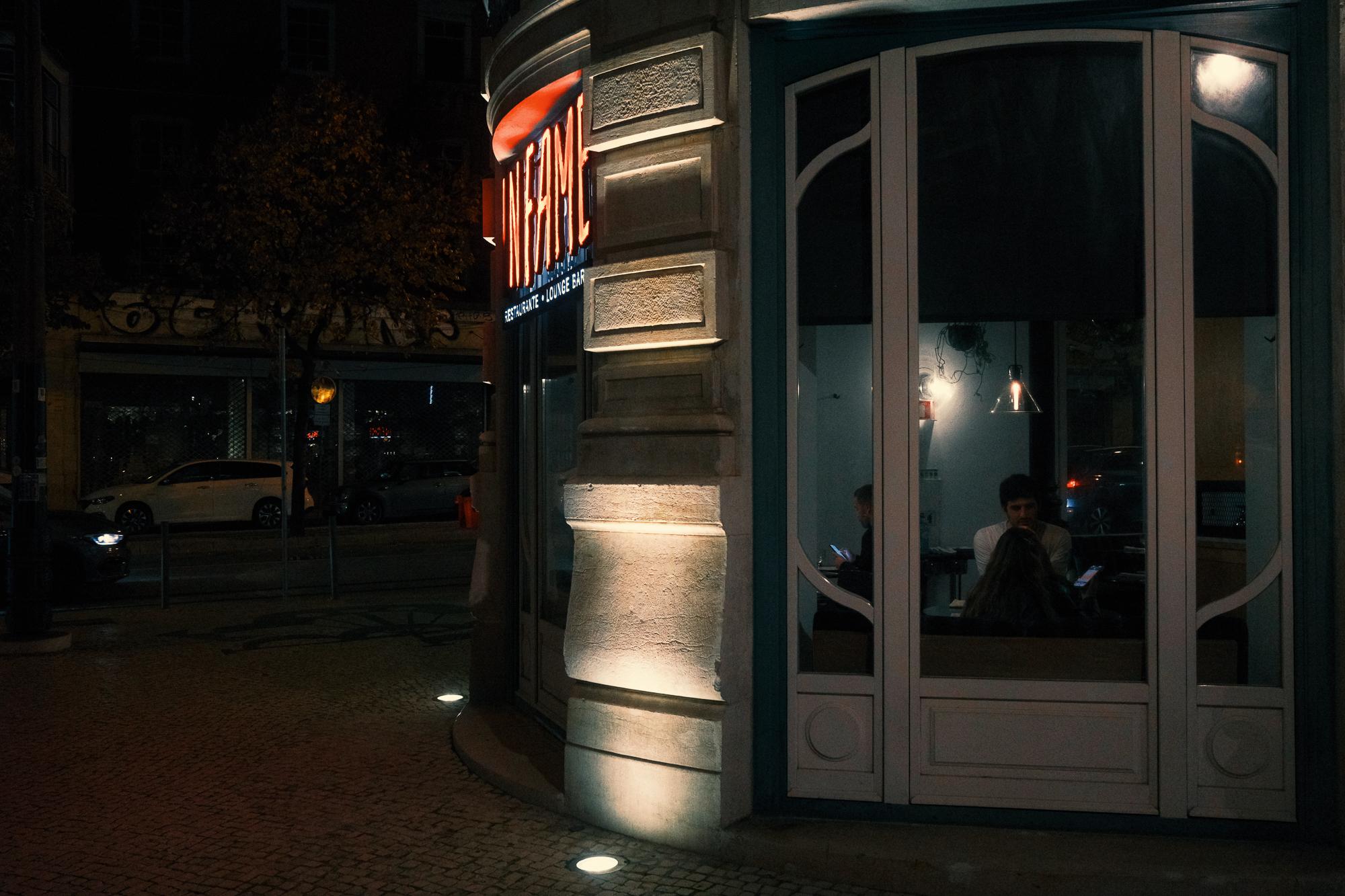 Nočný pohľad na roh budovy s osvetleným nápisom, ľudia sedia vo vnútri pri stole, jemné vonkajšie osvetlenie a autá na ulici.