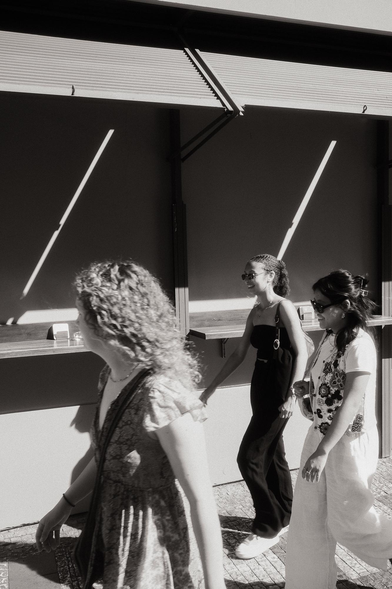 Tri ženy kráčajúce ulicou v letnom slnku. Na pozadí sú vidieť dlhé tiene odrážajúce sa na stene.