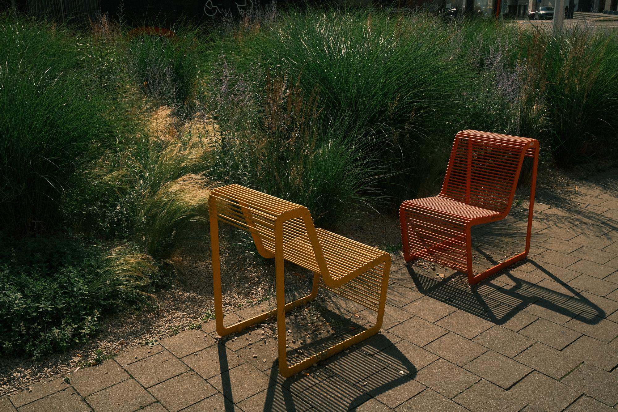 Dva farebné moderné kovové stoličky, jedna žltá a druhá oranžová, na dláždenom chodníku vedľa zelenej záhrady s vysokou trávou a kvetinami.