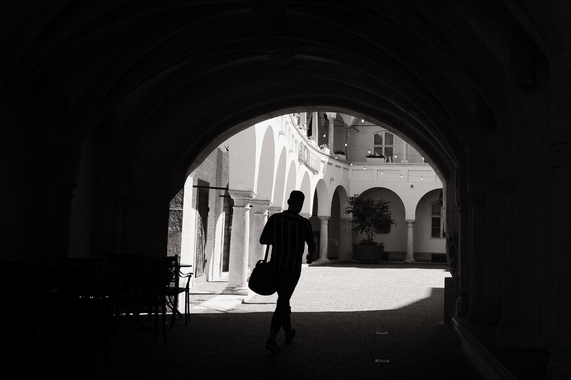 Čiernobiela fotografia zachytáva osobu kráčajúcu chodbou s elegantnými oblúkmi, ktorá vedie k slnečnému otvorenému dvoru s ozdobnými oblúkmi a pestro oblečenými rastlinami.