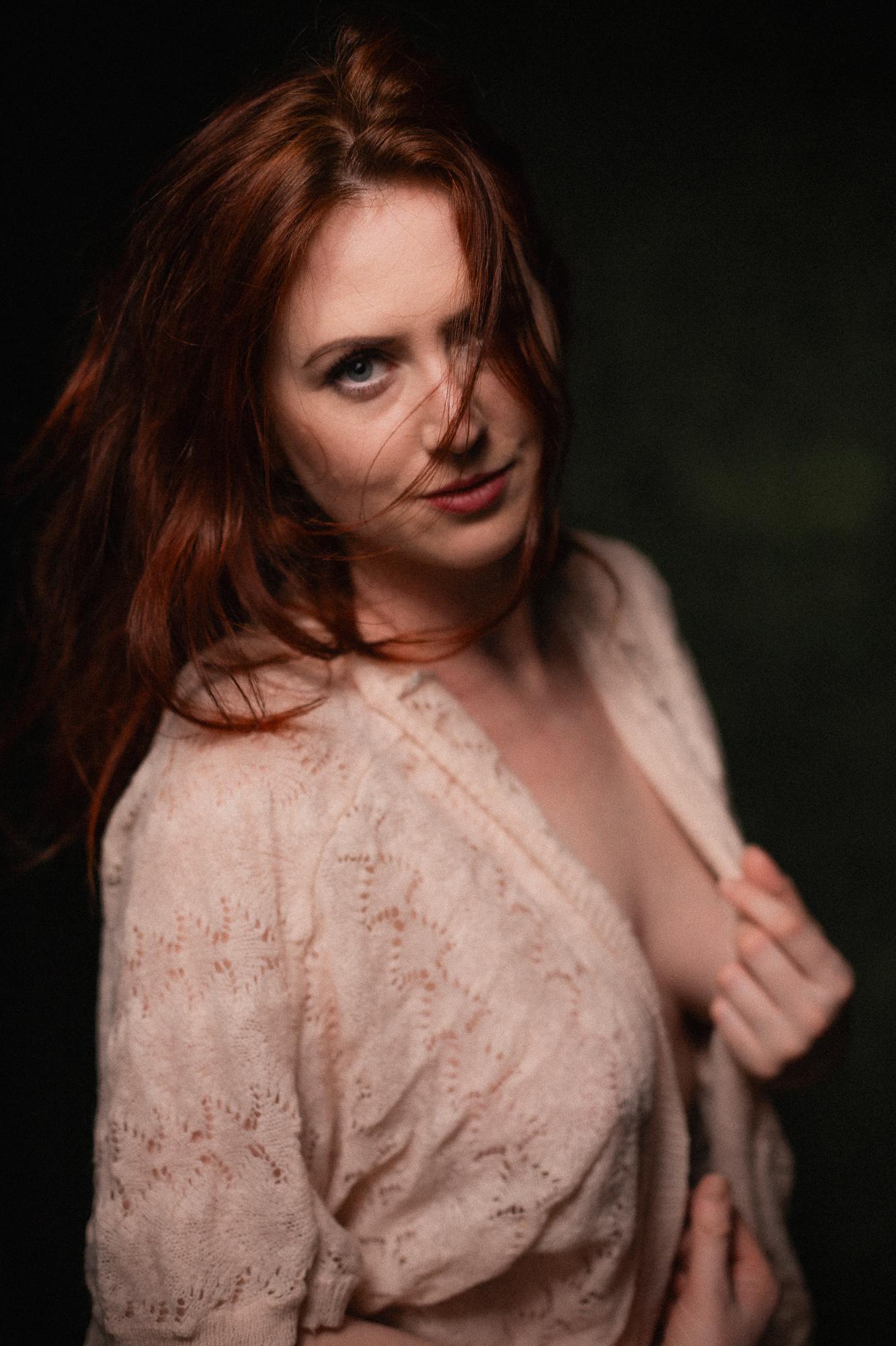 Portrét ženy s ryšavými vlasmi, ktorá sa jemne usmieva. Má na sebe béžový vzorovaný sveter a pozadie je tmavo zelené.