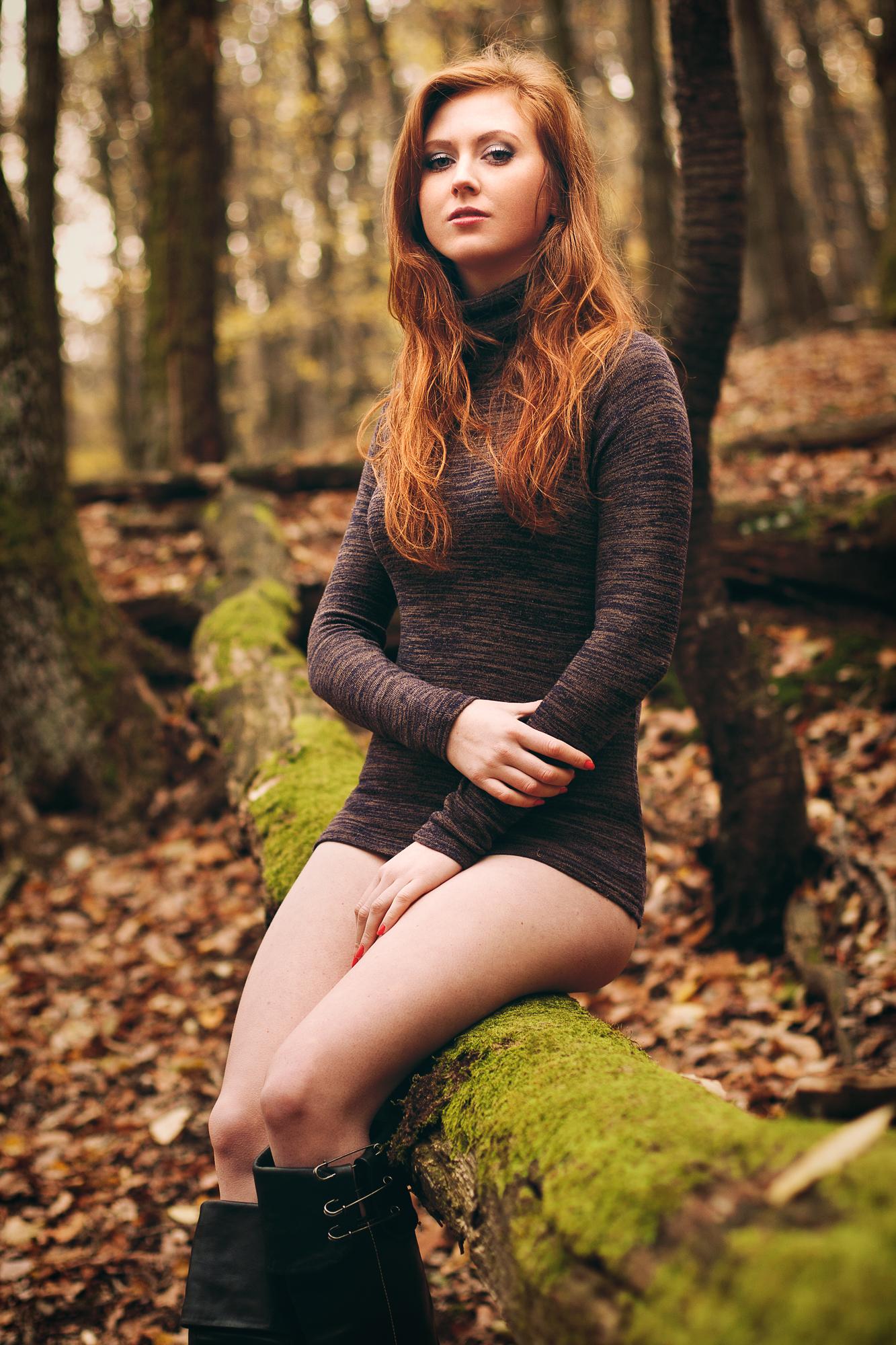 Portrét ženy s dlhými ohnivo ryšavými vlasmi, sediaca na machom pokrytom kmeni stromu v lese na jeseň. Na sebe má tmavošedý rolák a čierne čižmy.