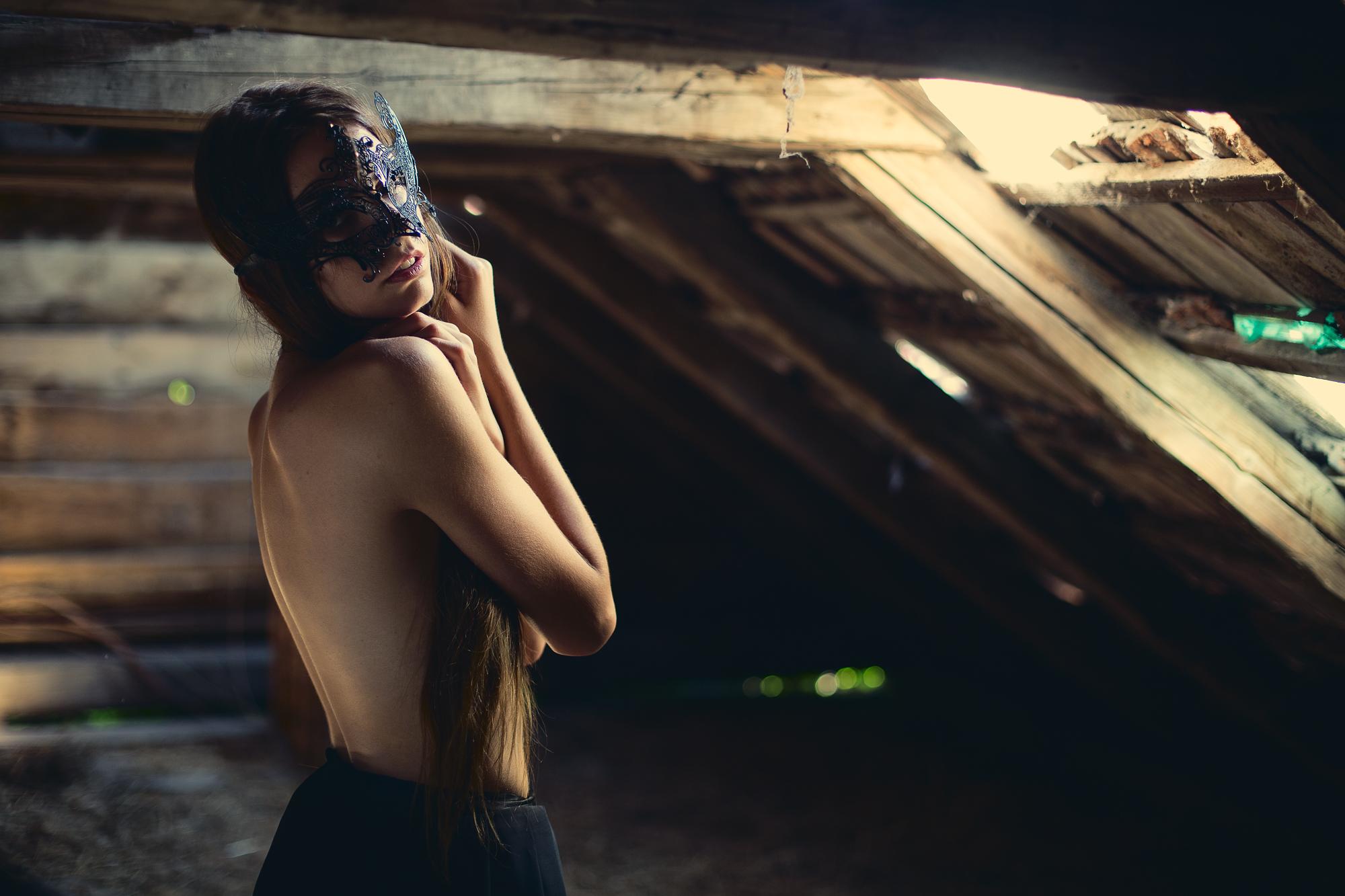 Žena s čiernou maskou na tvári, stojaca v strešnom podkroví osvetlená dierou v streche, oblečená iba do dlhej čiernej sukne a zahalená rukami.