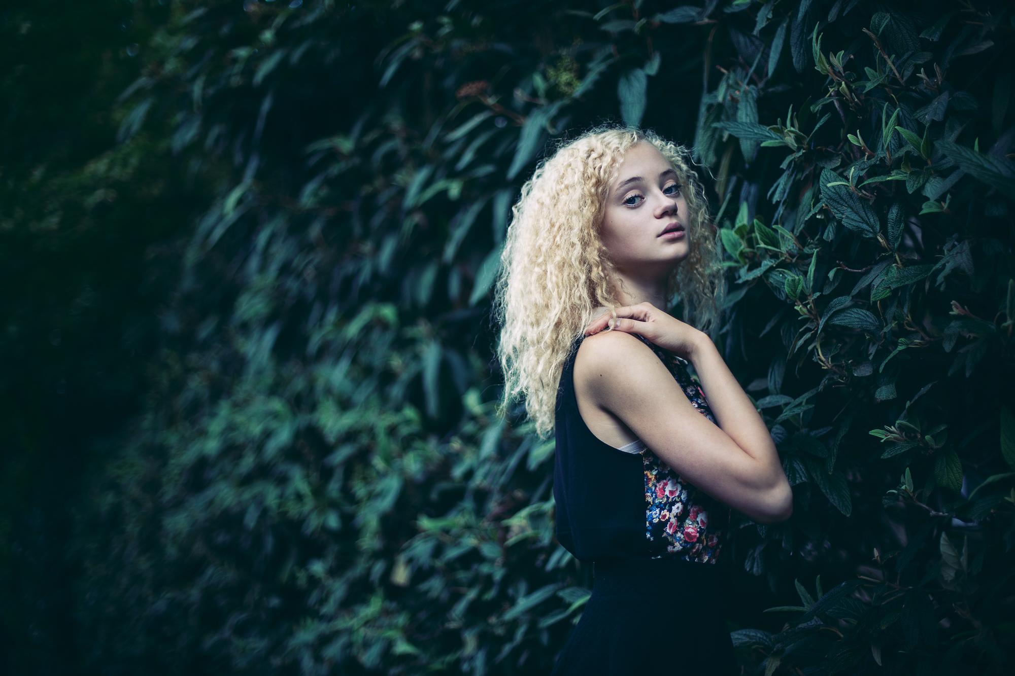 Portrét mladej ženy s blond kučeravými vlasmi, ktorá stojí pred zeleným pozadím lístia, oblečená v čiernom topu s kvetinovým vzorom.