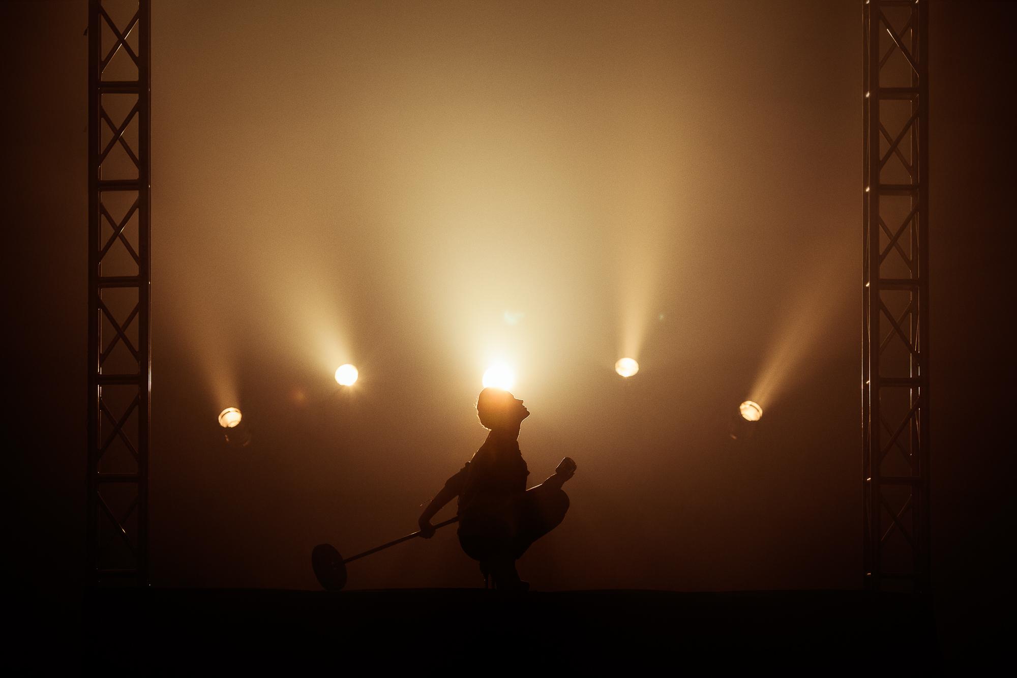 Silueta osoby s mikrofónom na javisku, osvetlená zozadu  lúčmi svetla z reflektorov usporiadaných v tvare vejáru v hmlistej atmosfére.
