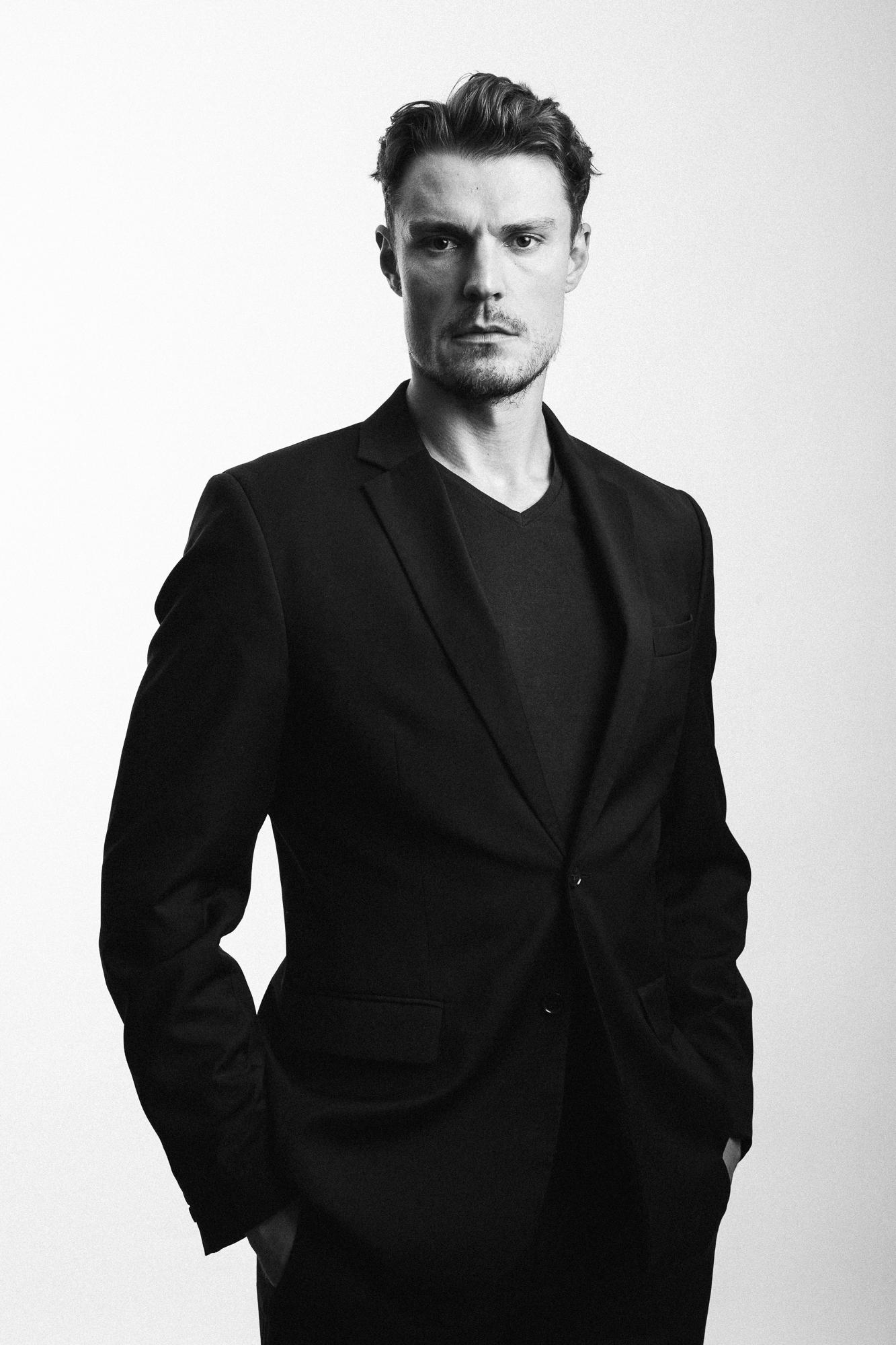 Čiernobiela fotografia muža v tmavom obleku stojaceho proti bielému pozadiu.