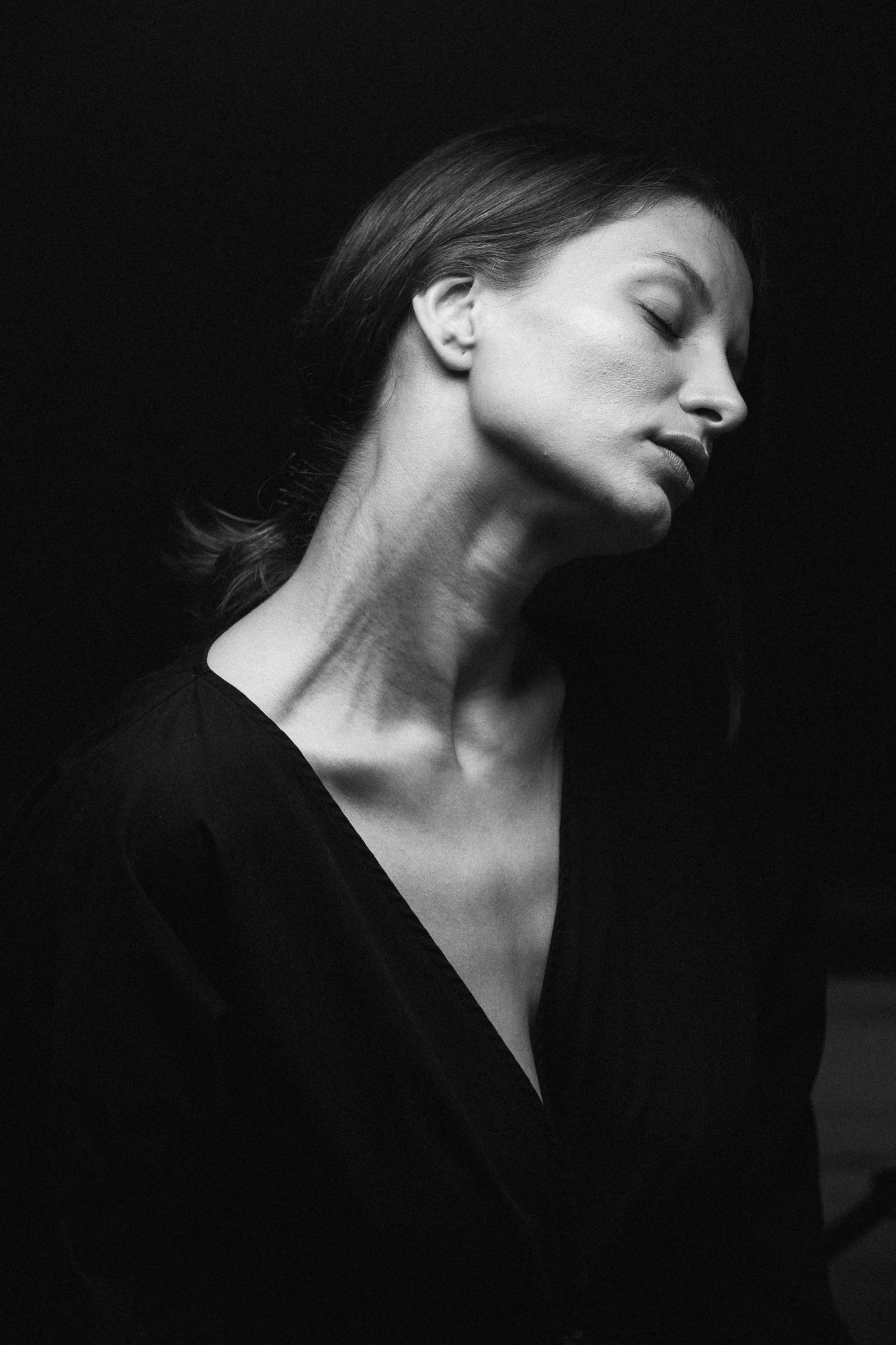 Čiernobiela fotografia ženy so sklopenou hlavou a zatvorenými očami, v čiernom oblečení na tmavom pozadí.