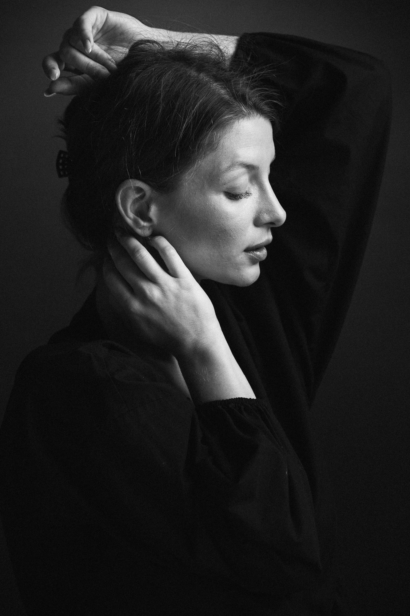 Čiernobiela fotografia ženy s jemne zatvorenými očami, rukou na krku a druhou rukou položenou na vlasoch. Má zamyslený a pokojný výraz.