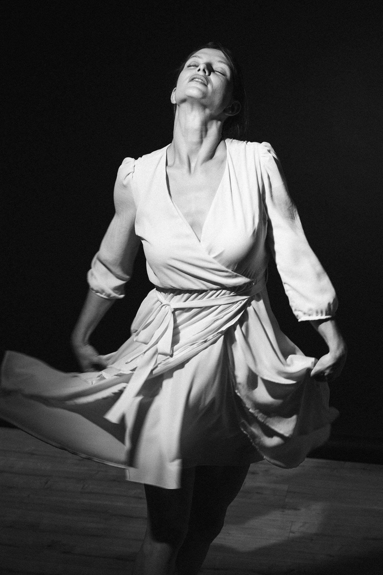 Čiernobiela fotografia ženy v bielych šatách, ktorá tancuje v silnom kontrastnom svetle.