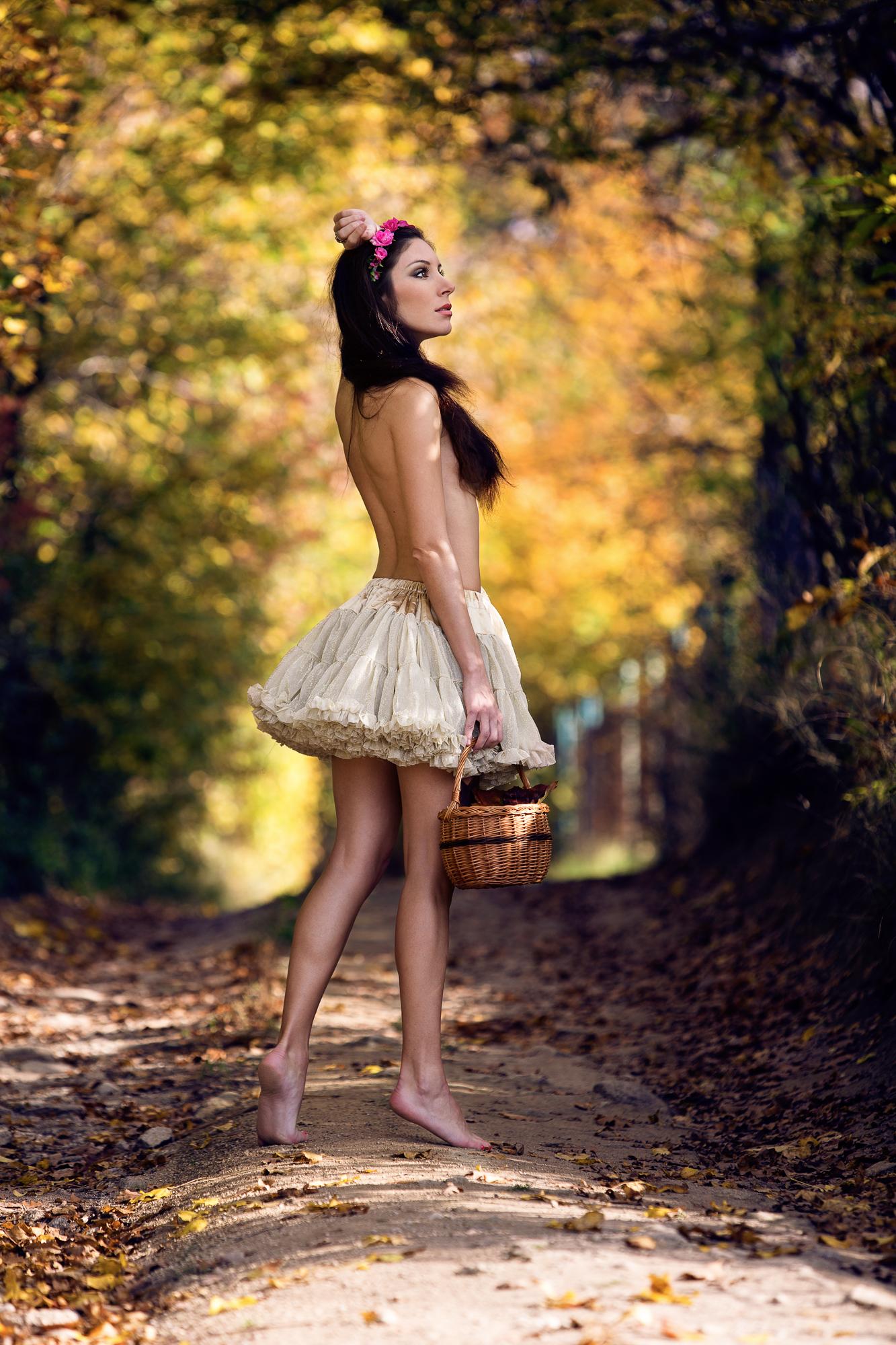 Farebná fotografia mladej ženy pripomínajúcej vílu, oblečenej iba do jemne bledej sukne držiacej prútený koš na lesnej ceste v jesennej scenérii s farebnými listami.