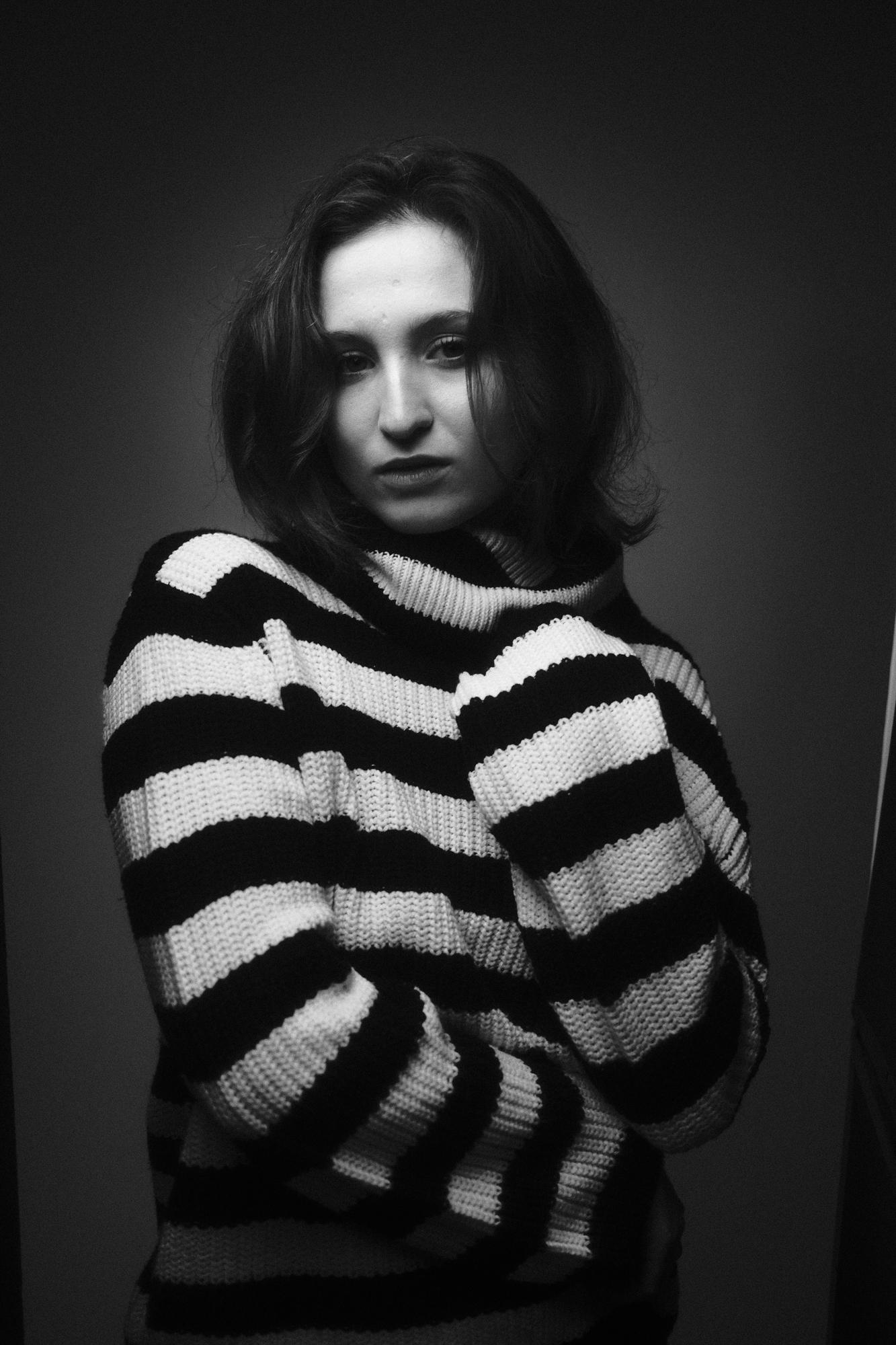 Čiernobiely portrét ženy v pruhovanom svetri, so zadumaným výrazom, s jemným svetlom na pozadí.