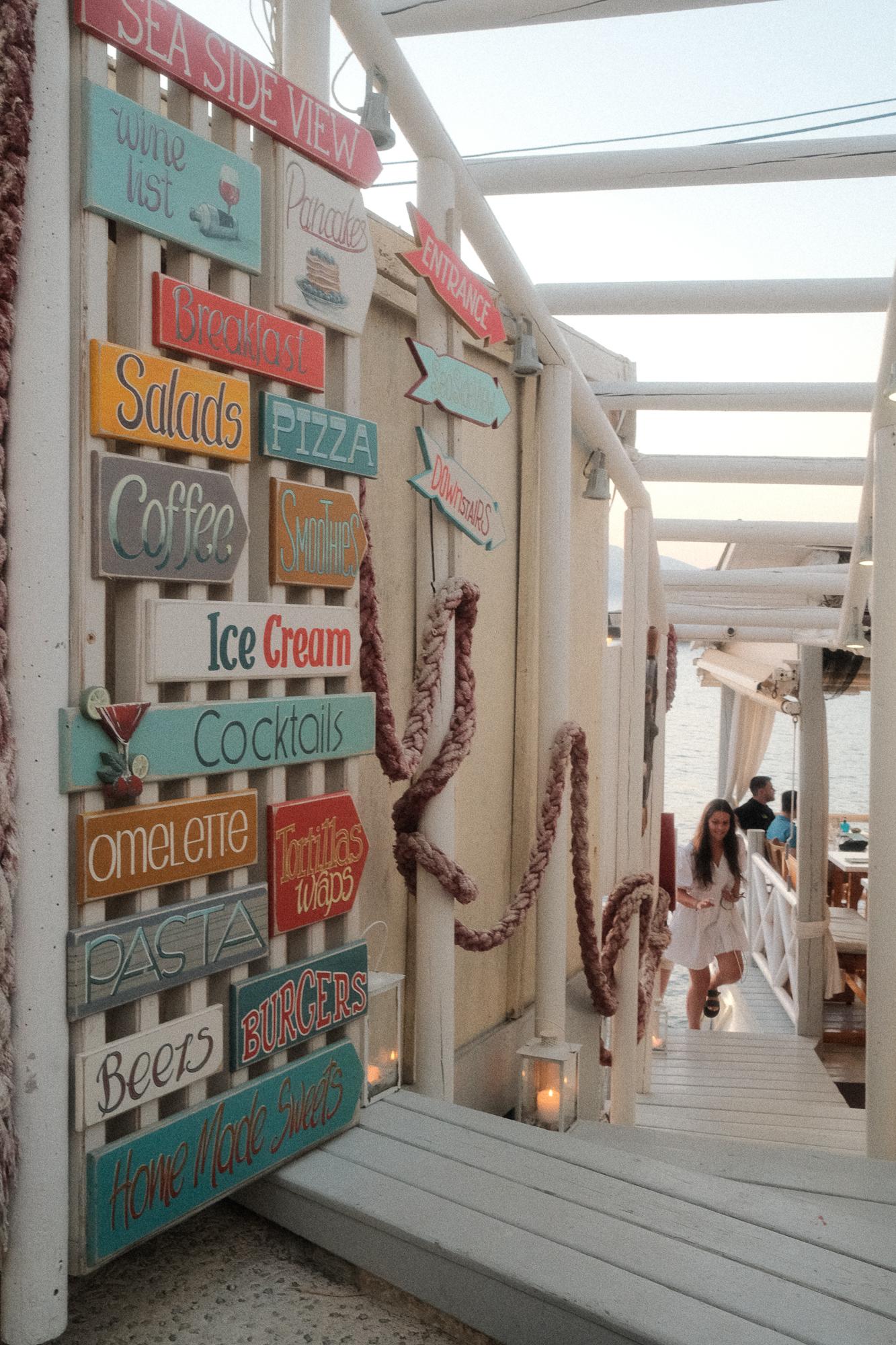 Farebné tabuľky označujúce ponuku jedál a nápojov na stene vonkajšej reštaurácie pri mori, vedľa schodiska, ktorým zostupuje žena v bielych šatách.