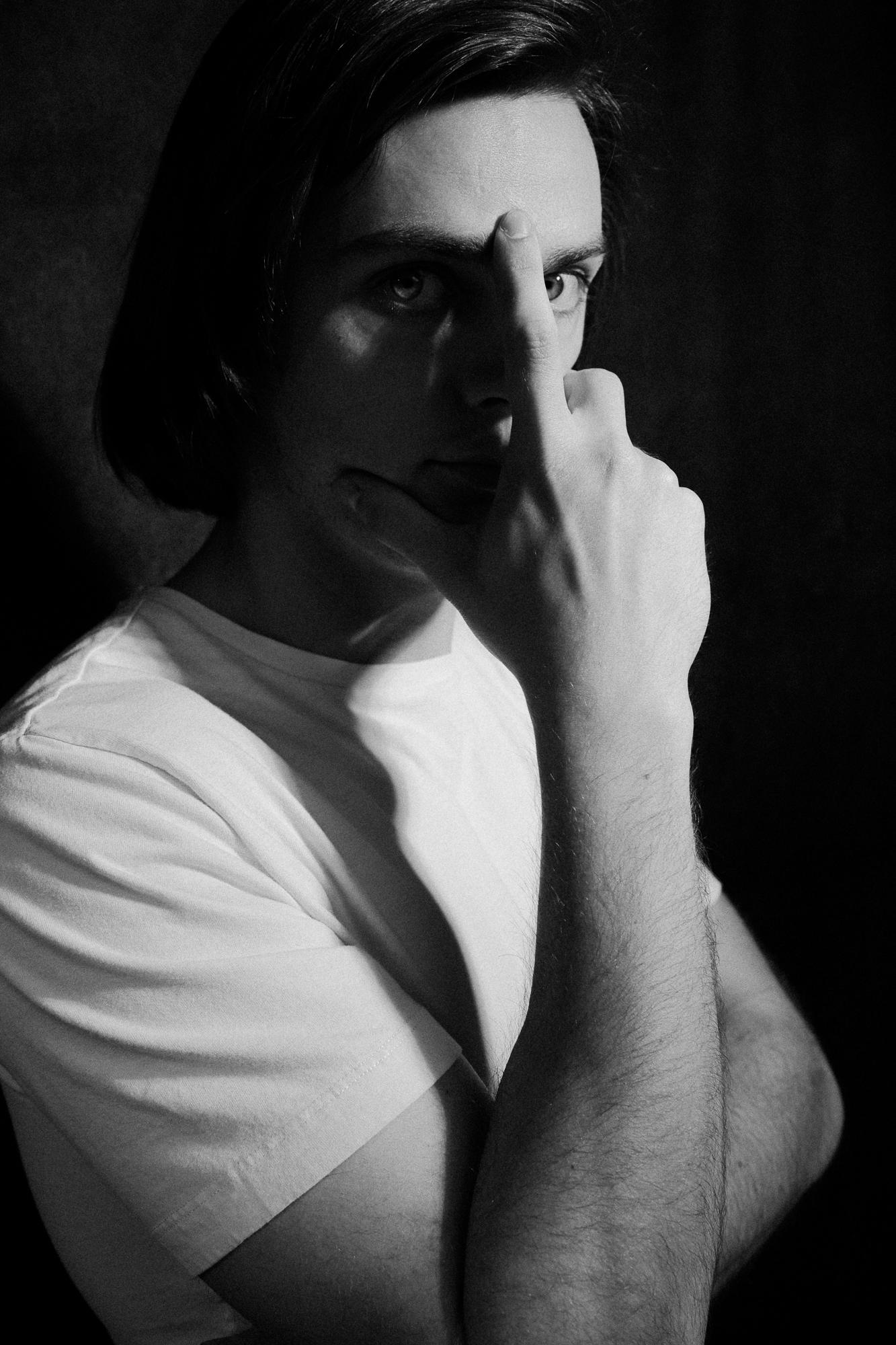 Čiernobiela fotografia muža v bielom tričku, s pravou rukou položenou na tvári a prstami tvarujúcimi rámovanie jeho oka, otočeného mierne na bok s priamym pohľadom do objektívu.