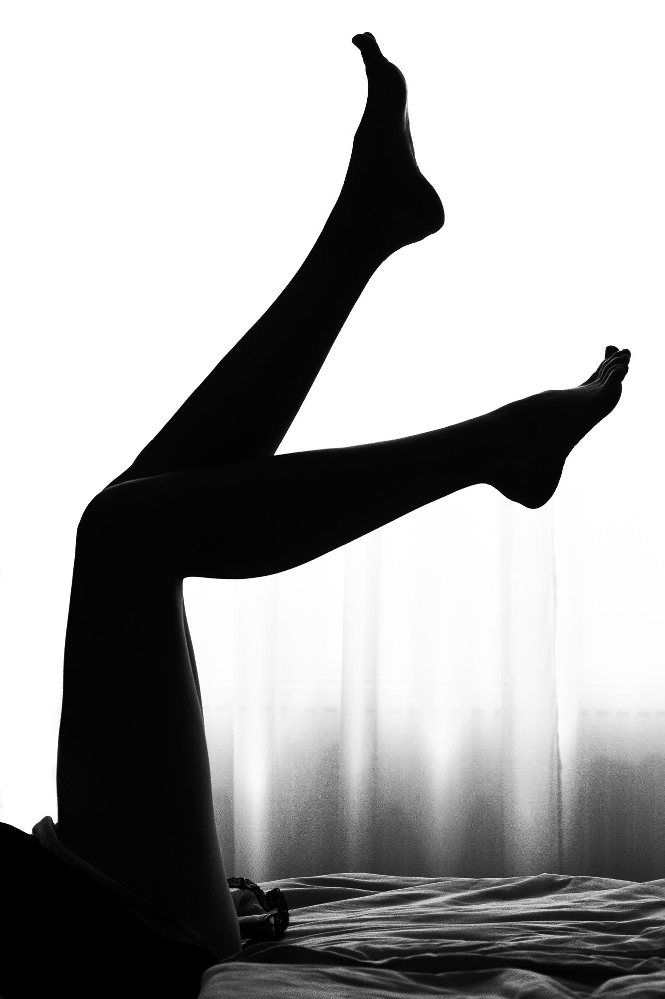 Čierno-biely umelecký akt zachytávajúci siluetu ženských nôh smerujúcich k stropu z postele pred presvetlenými závesmi. Praha.