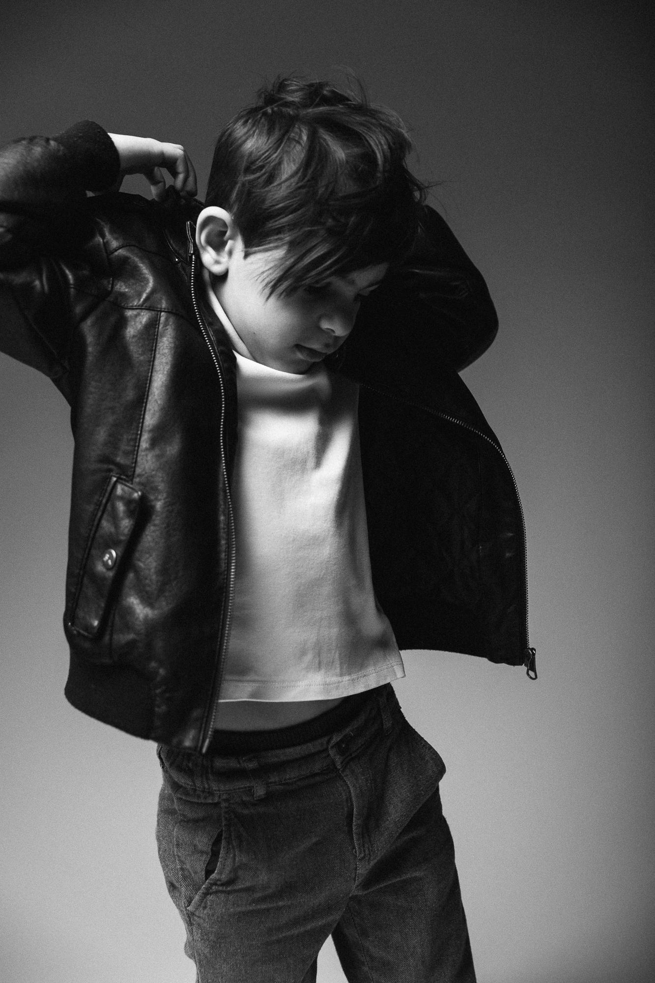 Čiernobiela fotografia malého chlapca, ktorý si oblieka bundu, biela košeľa a džínsy, neutrálne pozadie.