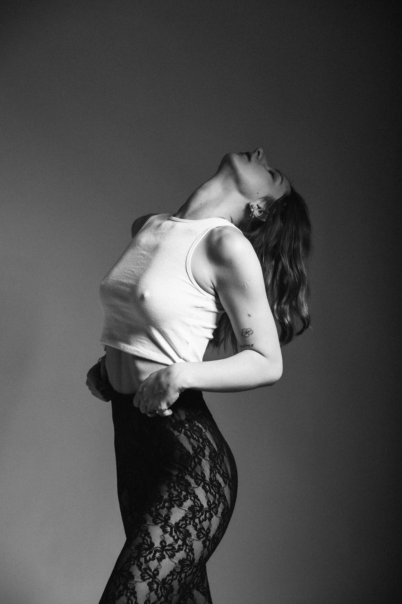 Čiernobiela fotografia ženy v bielom tielku a čipkovaných nohaviciach, zaklonená hlava a ruky položené na bedrách.