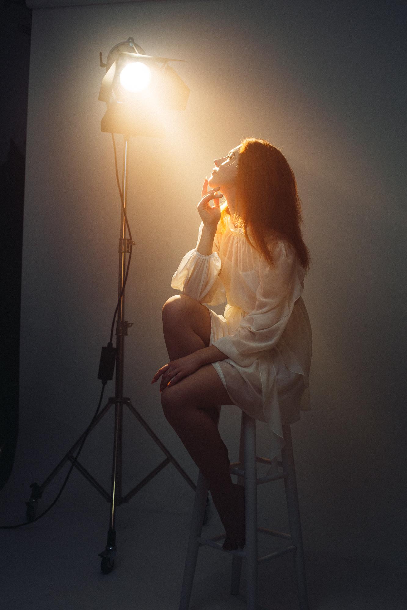 Žena sediaca na stoličke v štúdiu, oblečená v bielej blúzke, osvetlená silným svetlom filmárskym svetlom viditeľným v zábere.