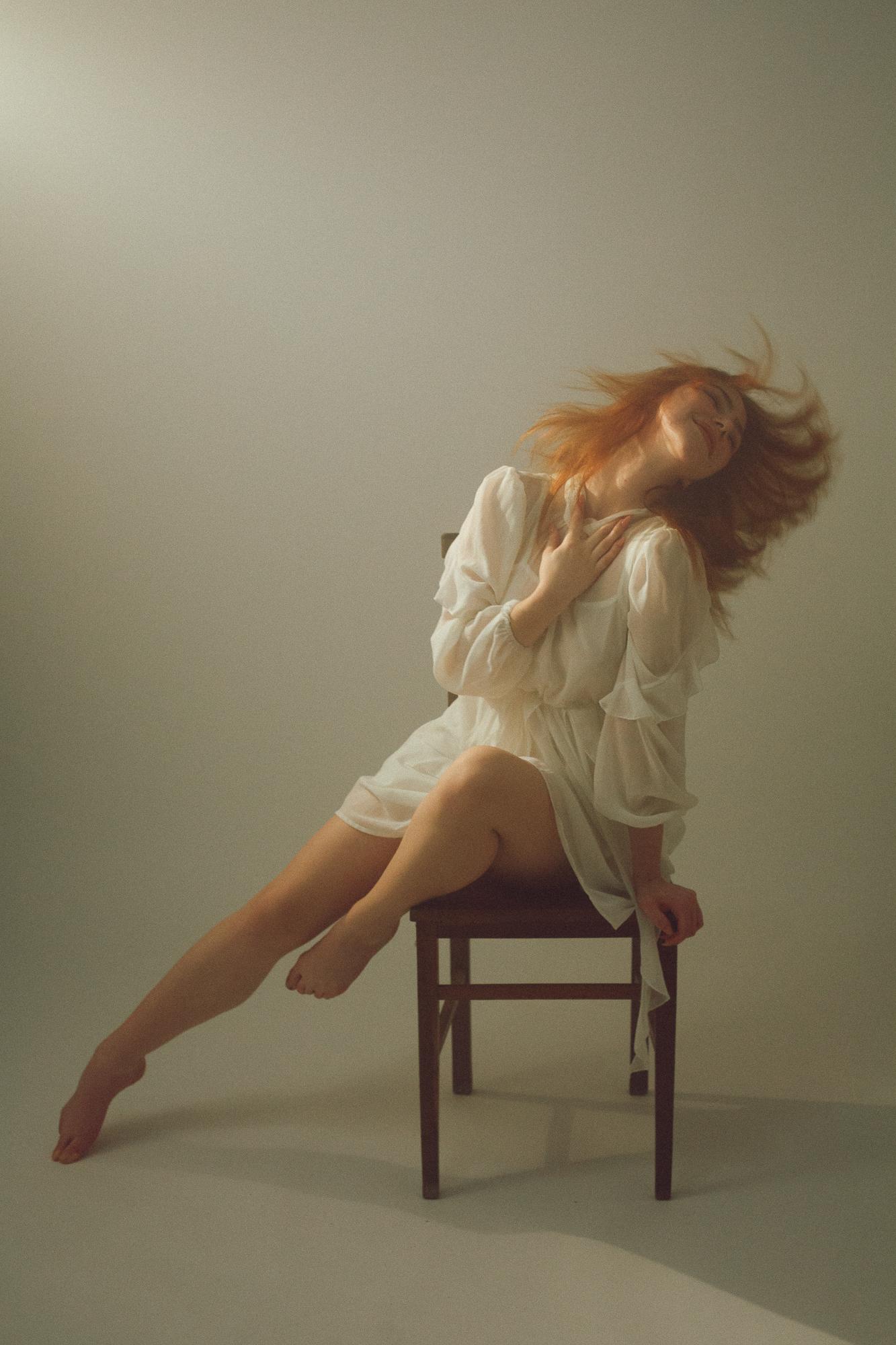 Žena s ryšavými vlasmi sediaca na stoličke, s hlavou zaklonenou dozadu, má na sebe biele splývavé šaty, jej vlasy sú v pohybe a jej výraz je uvoľnený a pokojný.