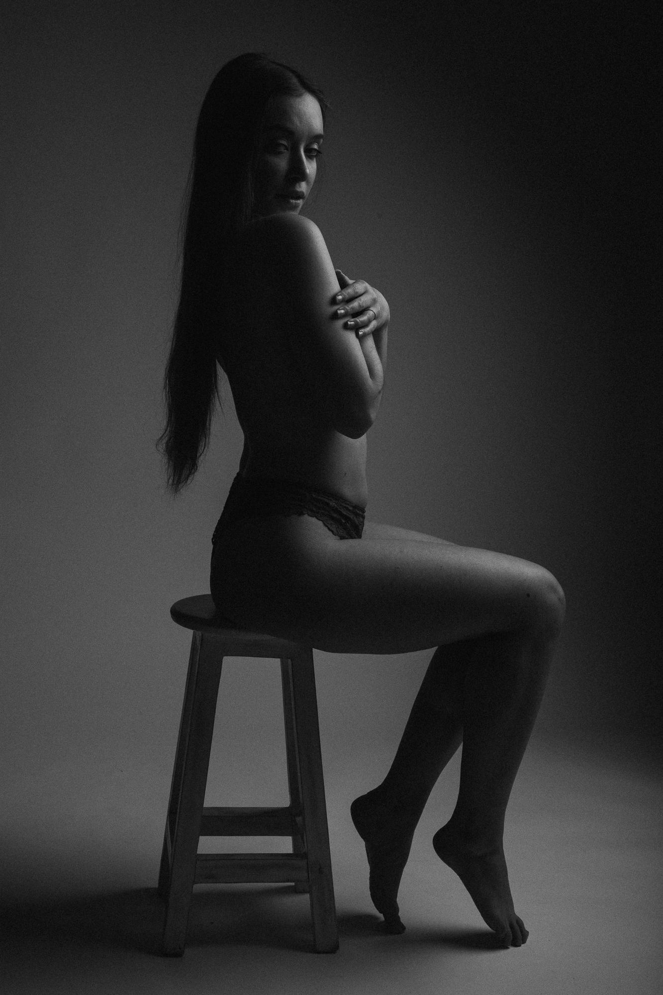 Čiernobiela fotografia mladej ženy sediacej na stoličke v spodnom prádle bez topu, s dlhými voľne padajúcimi vlasmi, nasvetlená mäkkým svetlom z boku, s rukou si zahalujúca hruď.
