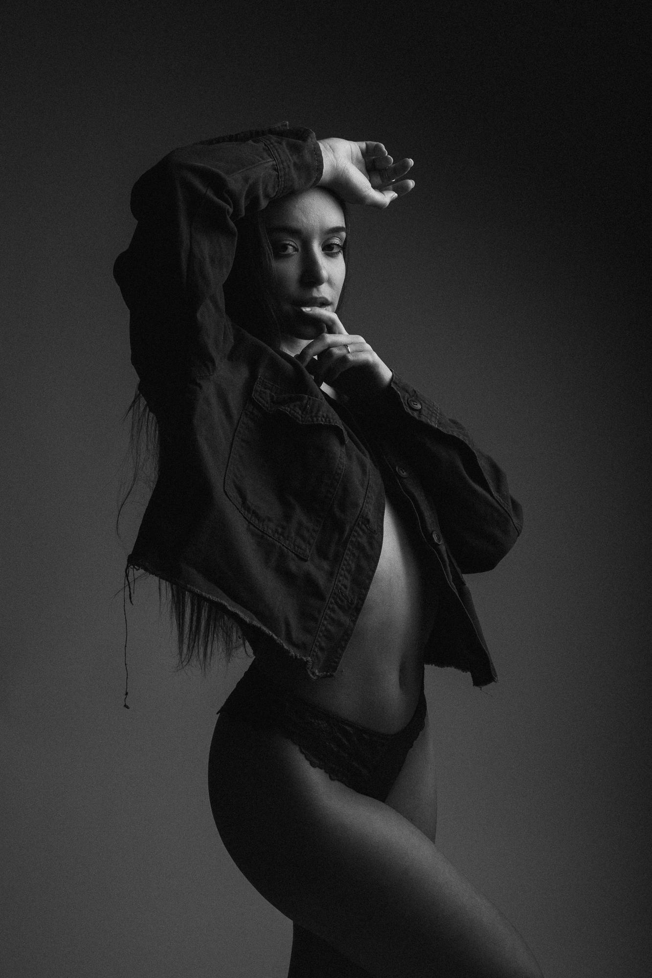 Čiernobiela fotografia modelky v džínsovej bunde a spodnej bielizni. Modelka má ruku nad hlavou a jemne sa dotýka tváre. Svetlo vytvára jemné tiene na tmavom pozadí.