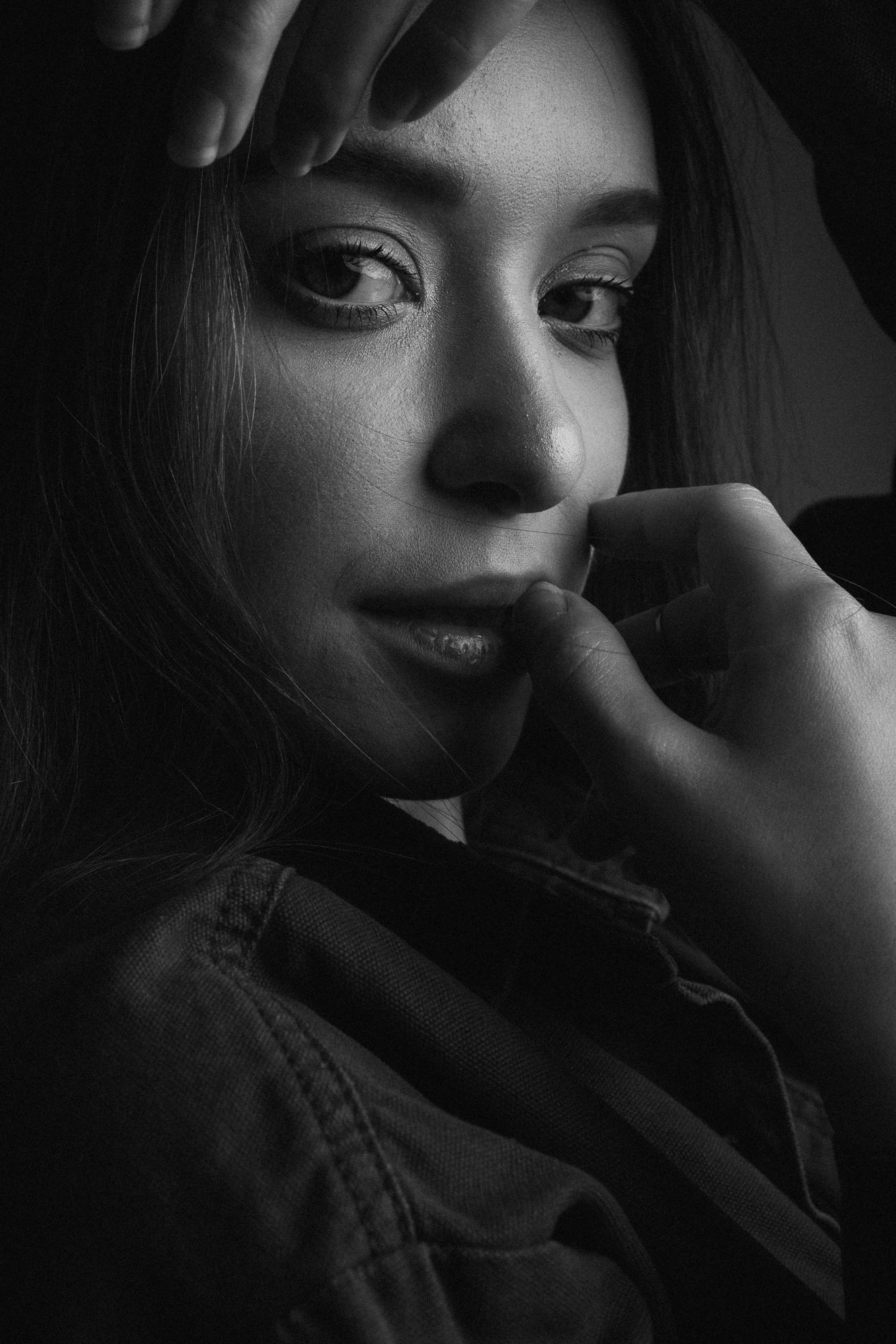 Čiernobiely portrét ženy s jemným svetlom zvýrazňujúcim jej oči. Jedna ruka sa jemne dotýka úst, druhá rámuje oči.