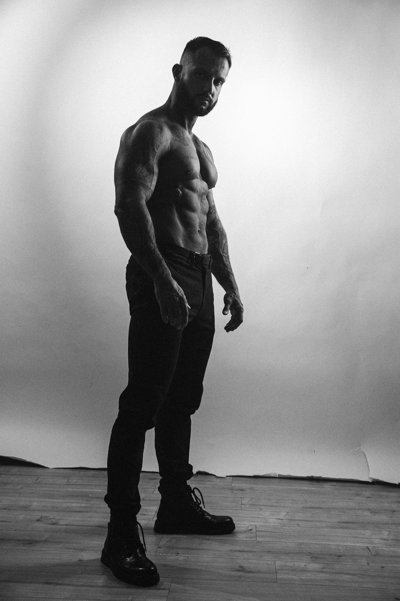 Kontrastný čiernobiely portrét muža s vyrysovanými svalmi stojaceho pred potrhaným bielym papierovým pozadím v ateliéri.