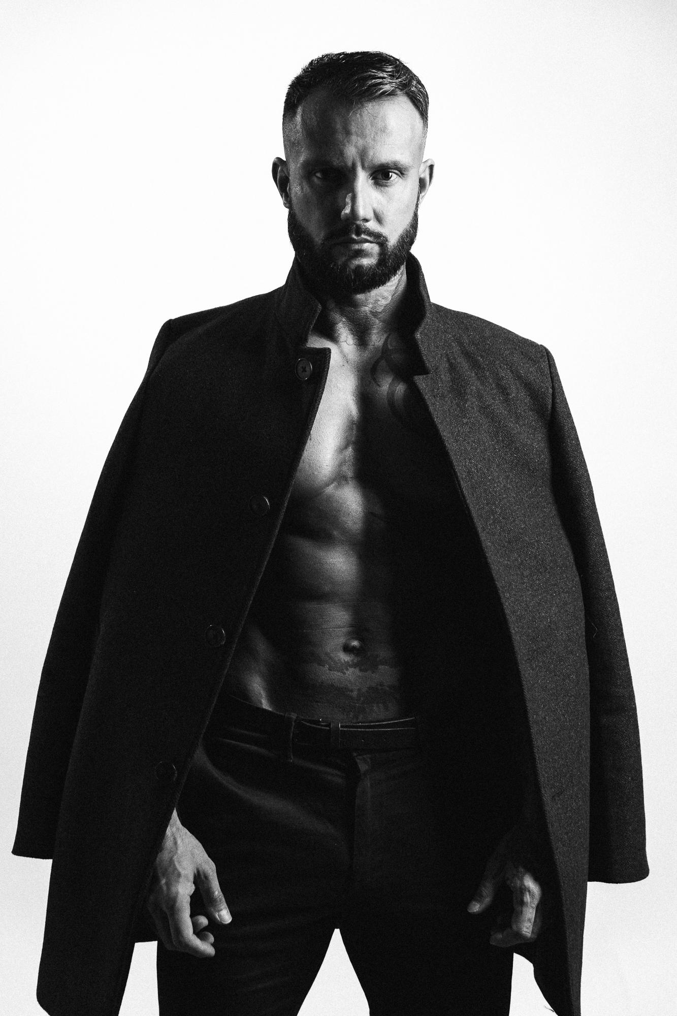 Muž v čiernom rozopnutom kabáte stojí proti svetlému pozadiu, odhaľujúci vypracovanú hruď.