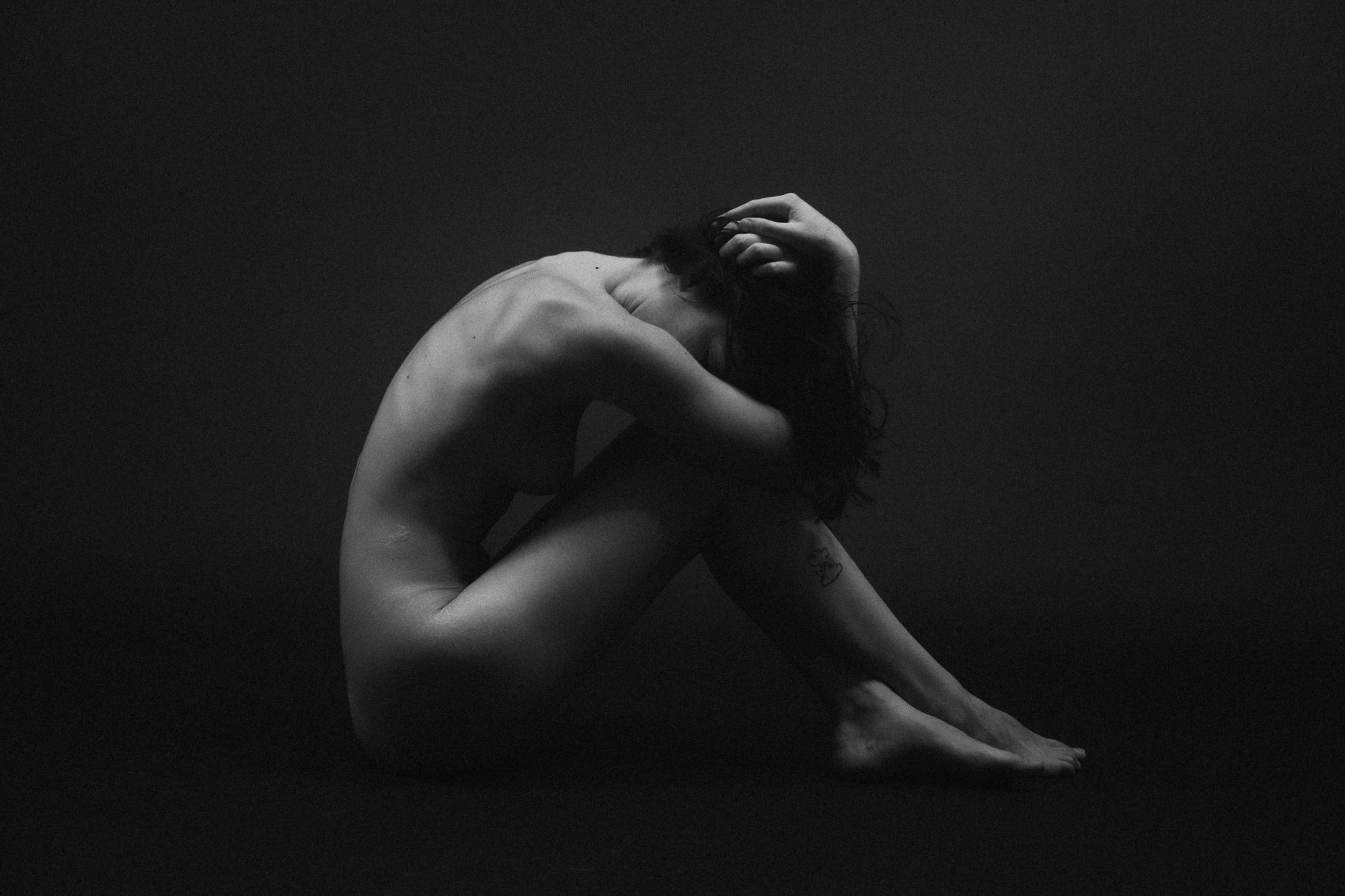 Čierno-biely umelecký akt. Mladá žena sedí na zemi, zľahka zahalená vlastnými rukami a vlasmi, v expresívnej pozícii. Ateliér Pezink.