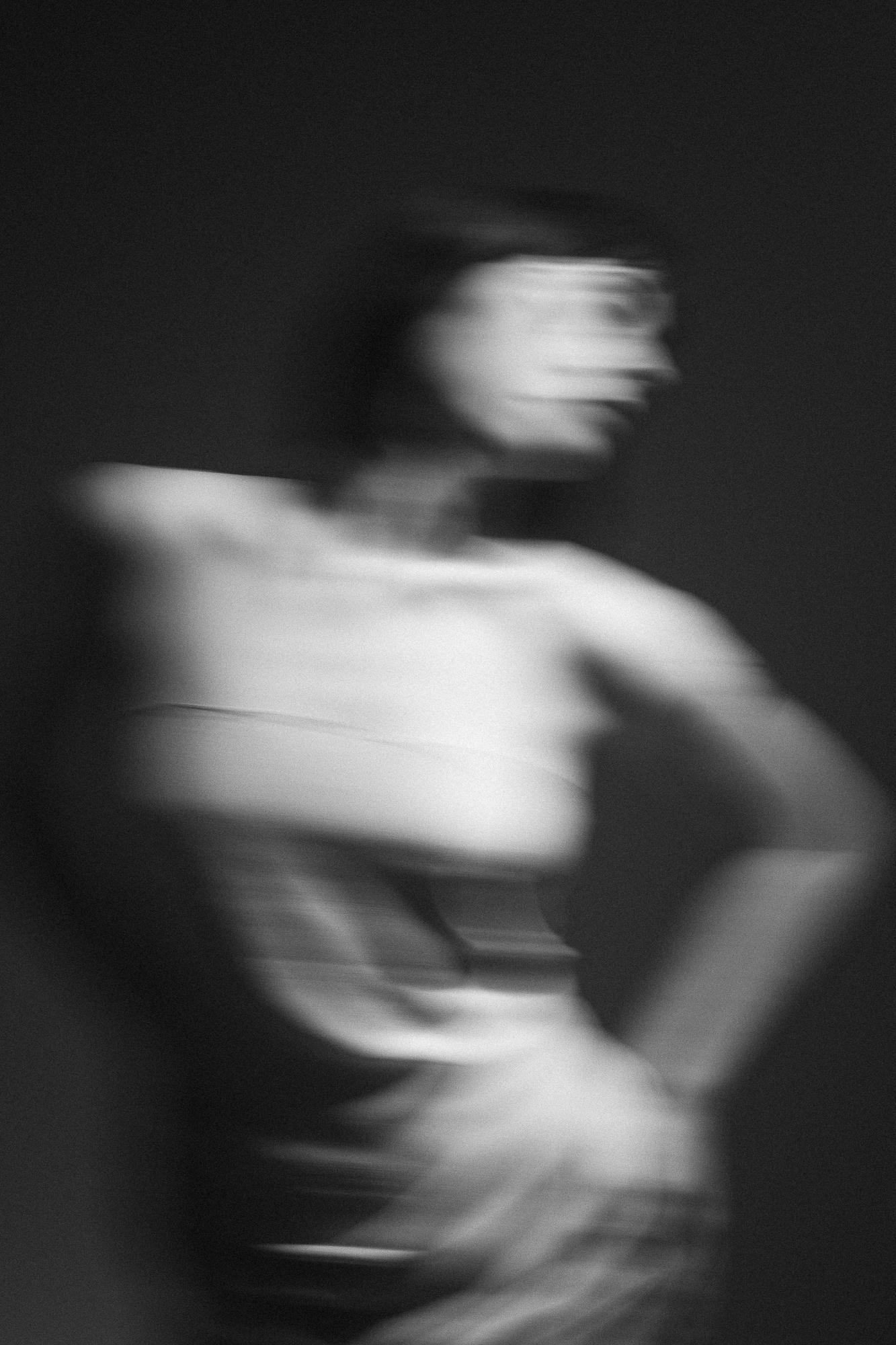 Čierno-biela rozmazaná fotografia zobrazujúca postavu ženy v pohybe zahalenej do bledej látky, s rukami na bokoch, proti tmavému pozadiu.